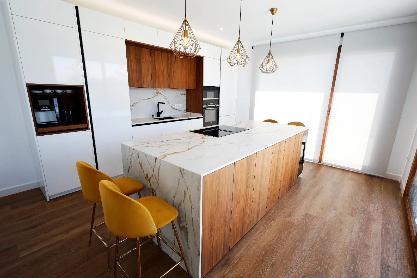 Modern kitchen with white and wooden cabinetry, marble countertop island, yellow bar stools, built-in oven, and large windows with blinds.