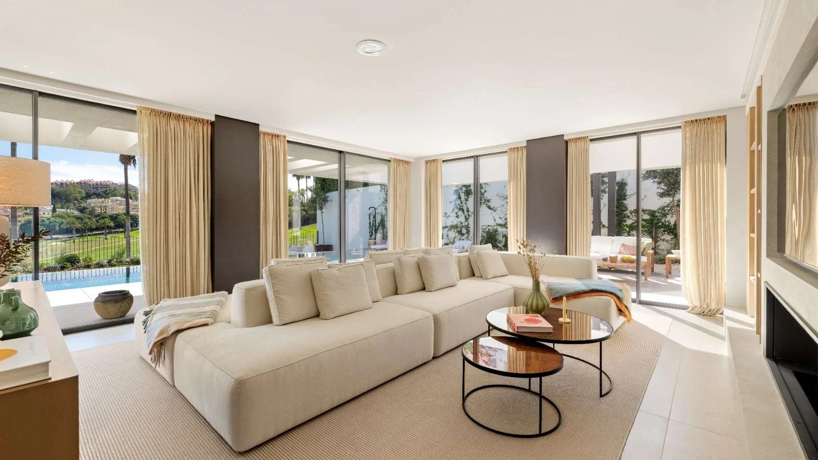 Bright living room with large beige sectional sofa, big windows with beige curtains, sliding glass doors leading to an outdoor patio, and a view of a backyard with a pool and greenery.
