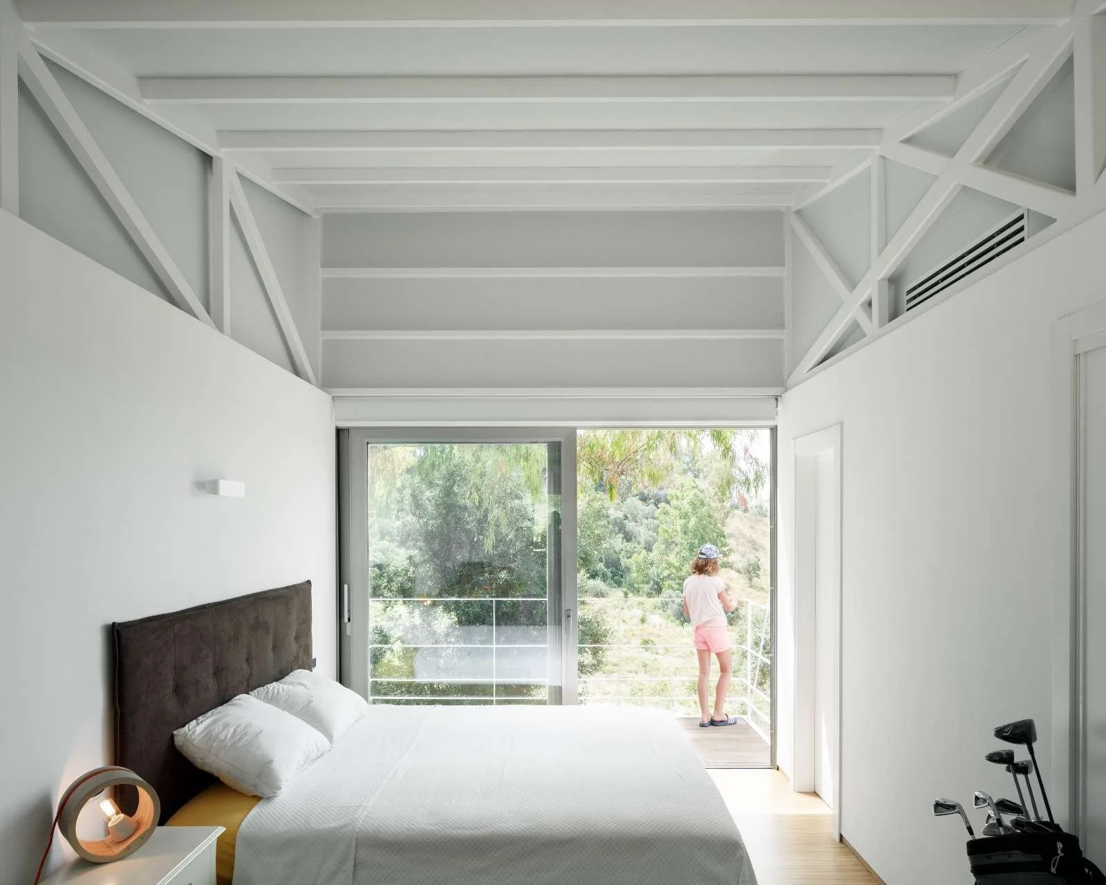 Modern bedroom with a large bed, a bedside lamp, and a view of a balcony where a woman in pink shorts and a white shirt is standing, looking at the trees outside.