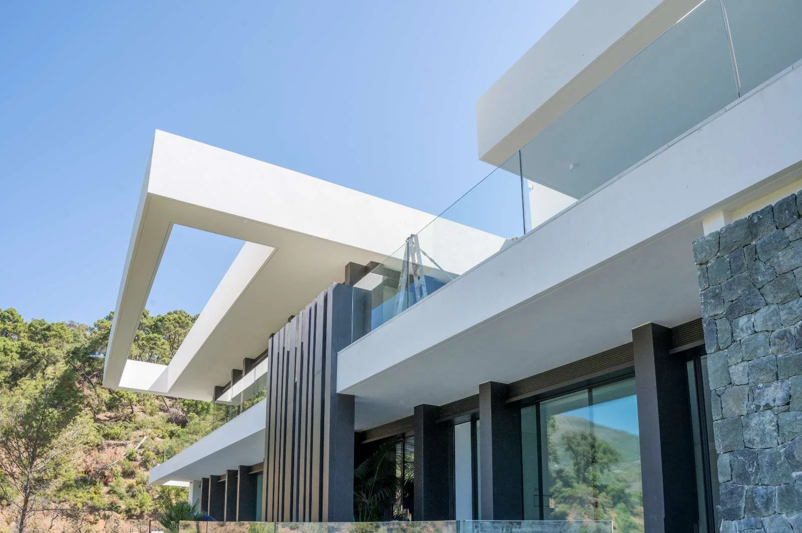 Modern house with white angular roof, glass balcony, stone wall, and large windows, surrounded by greenery under a clear blue sky.