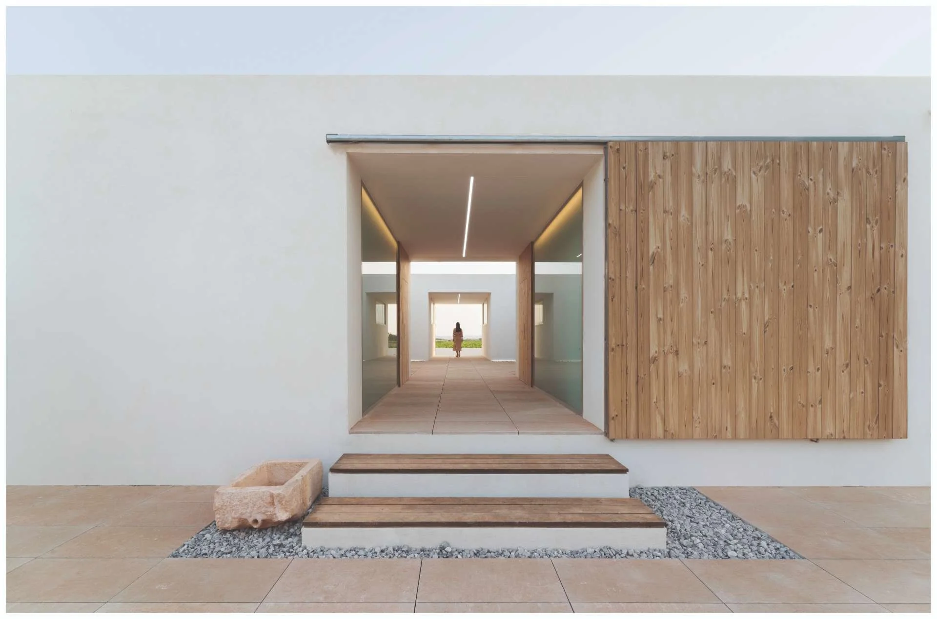 Modern minimalist architecture with white walls, wooden sliding door, stone steps, and a woman standing at the end of a corridor.