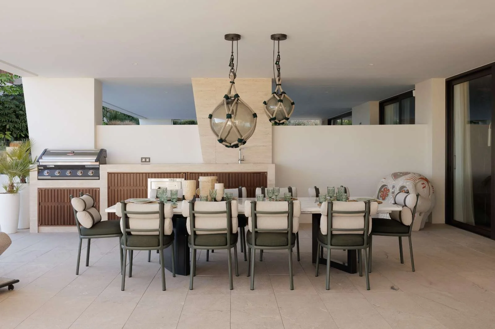 Outdoor dining area with a large rectangular table, multiple chairs, two hanging glass pendant lights, a built-in barbecue grill, and decorative items, with a sliding glass door on the side.