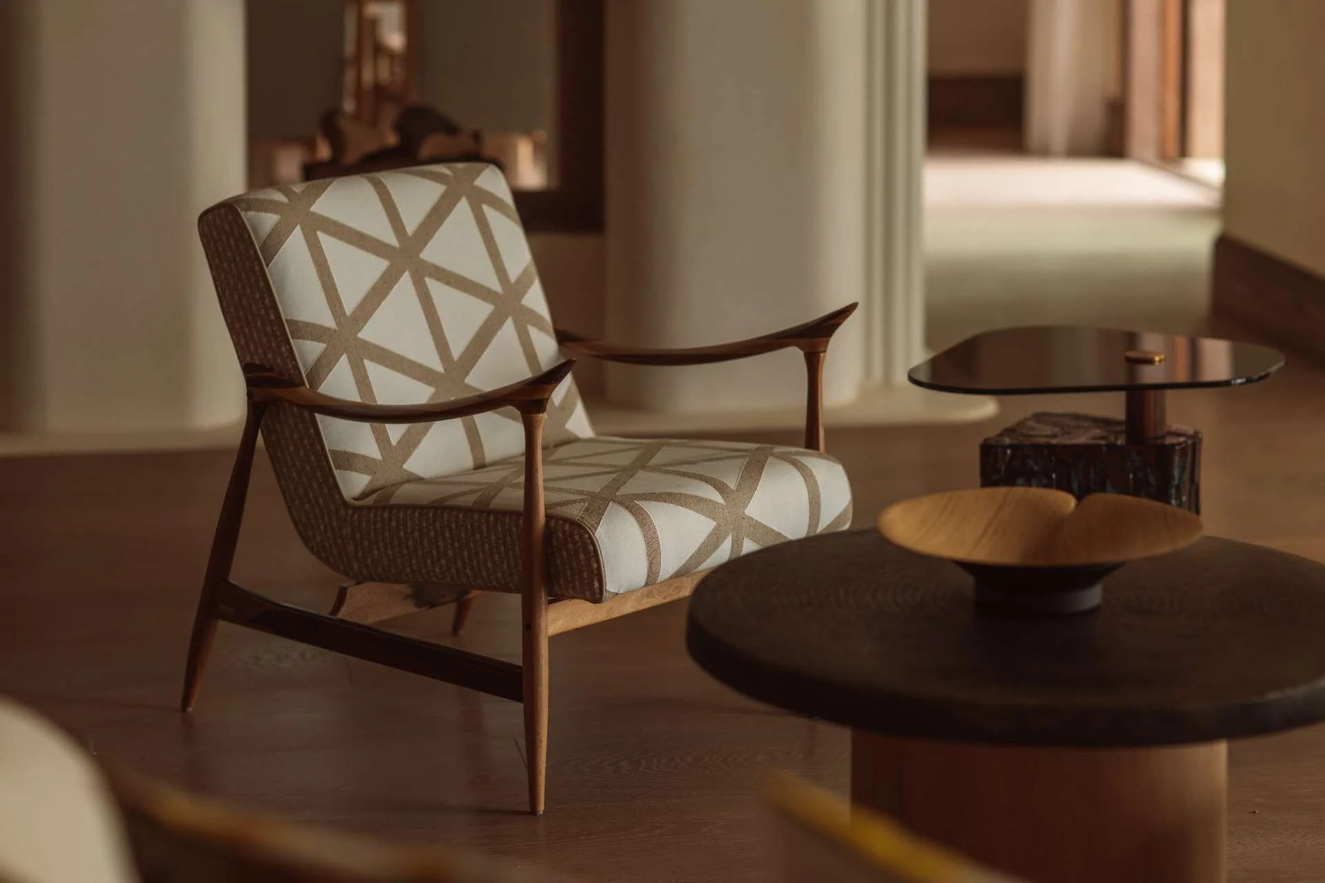 A mid-century modern armchair with a beige and white geometric patterned cushion, situated in a warmly lit room with wooden furniture and a glimpse of a hallway in the background.