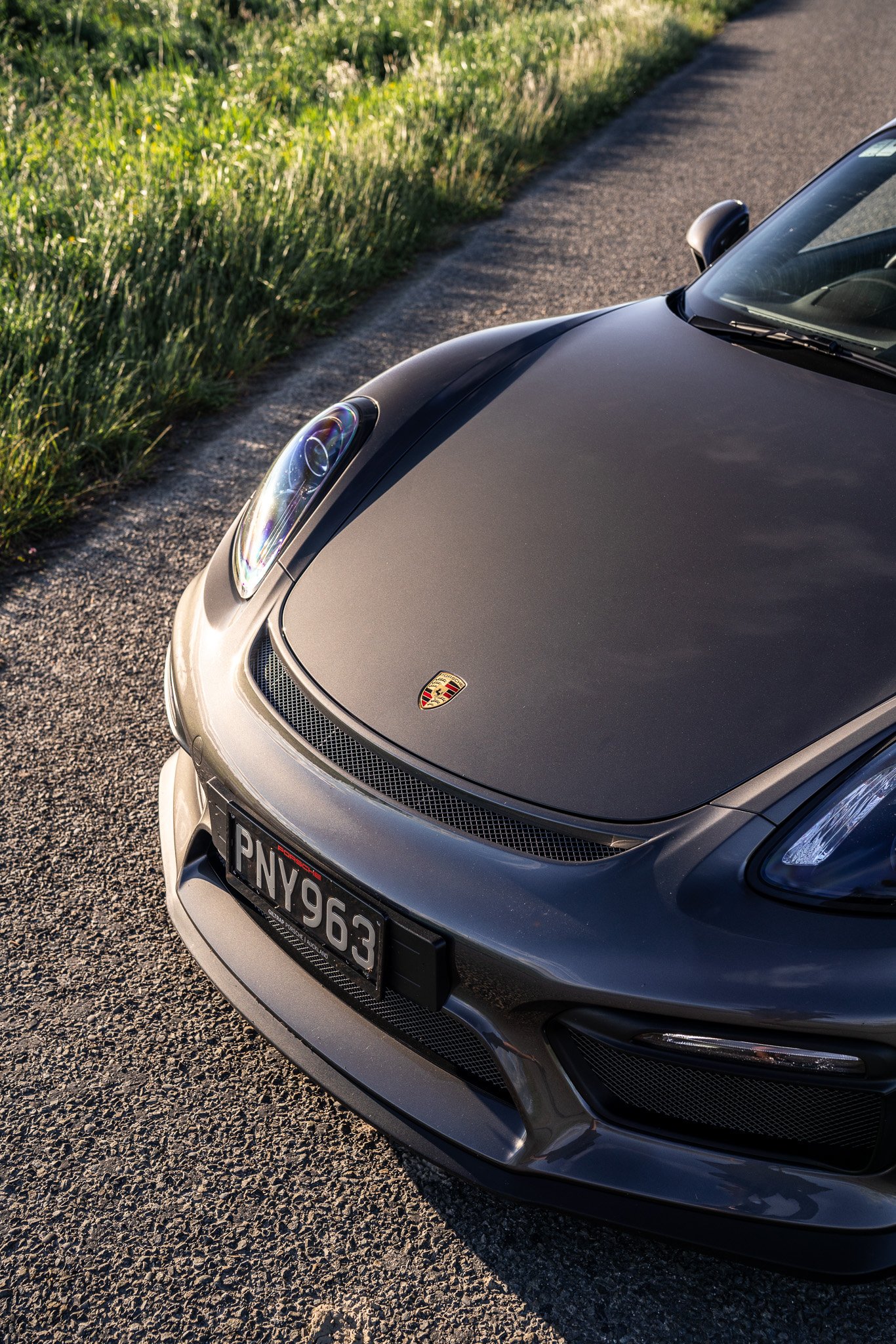 Close-up view of the front of a gray Porsche sports car parked on the side of a rural road with grass and trees in the background.