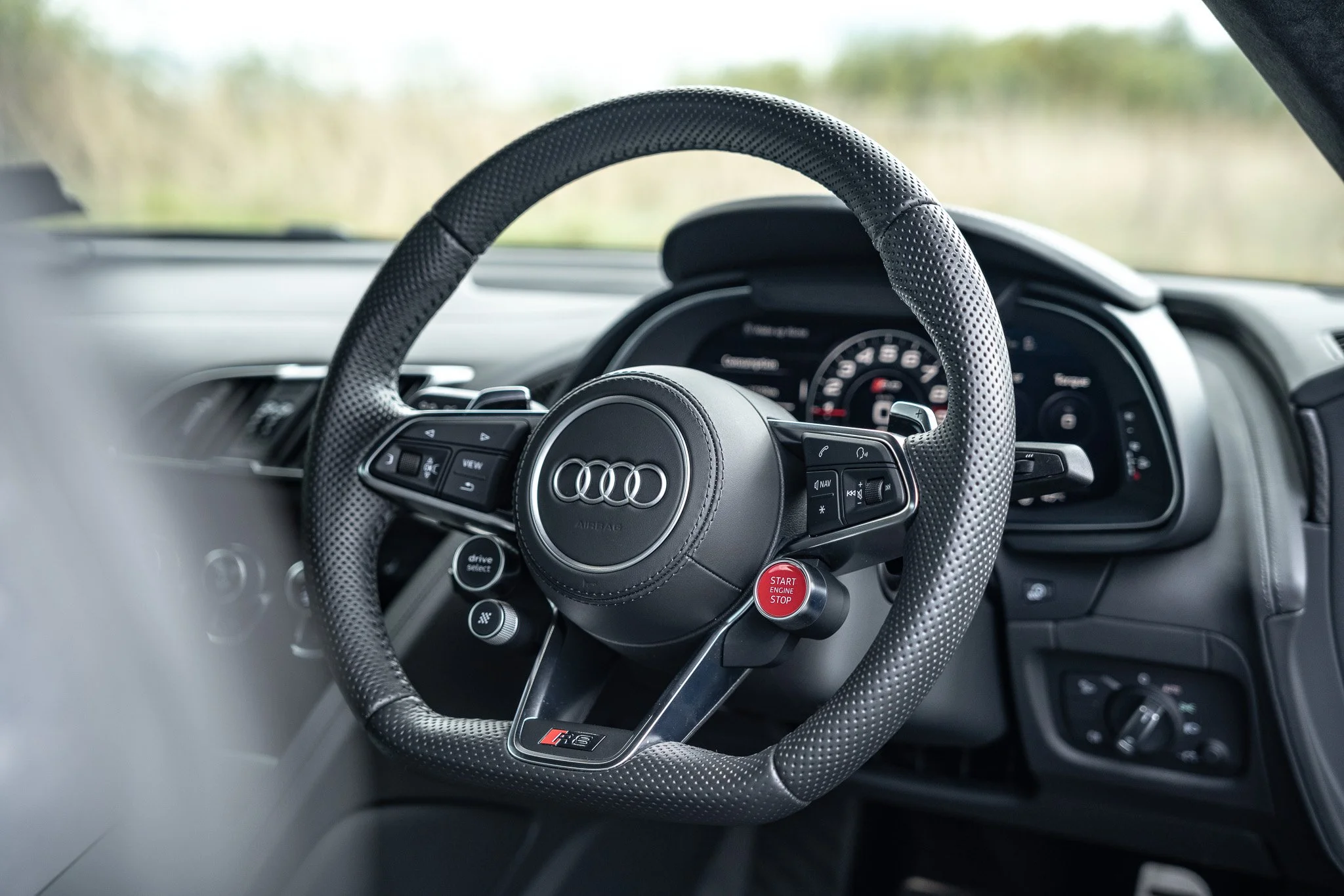 Interior of an Audi car, showing a steering wheel with Audi logo, dashboard with speedometer, and start/stop button.