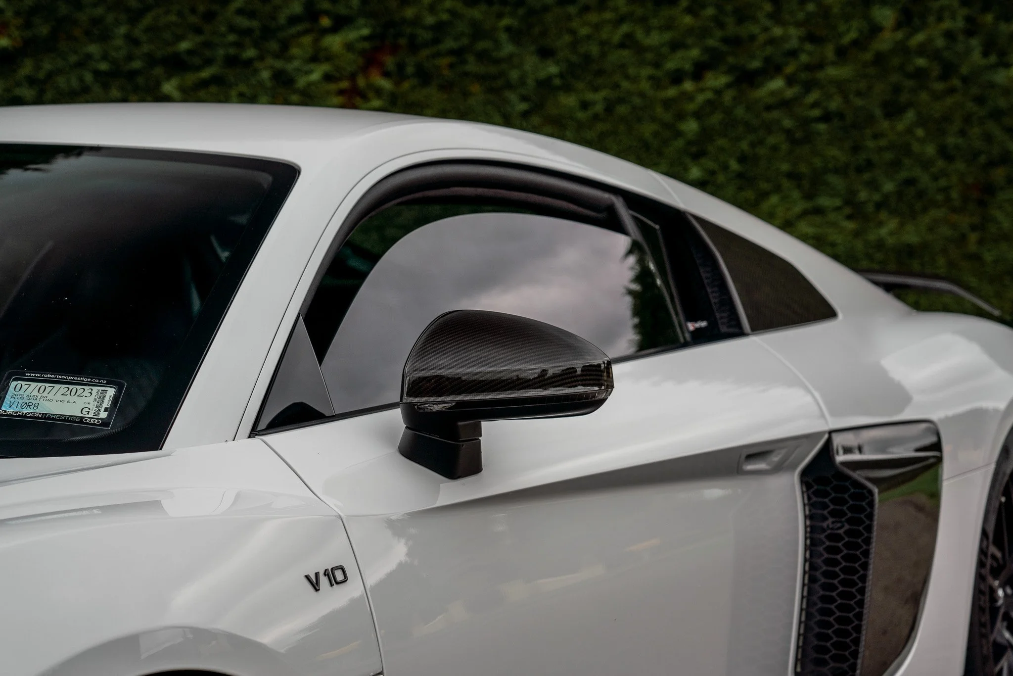 Close-up of a white luxury sports car with a carbon fiber side mirror and V10 badge, showing part of the windshield and side vent, set against a background of greenery and cloudy sky reflection.