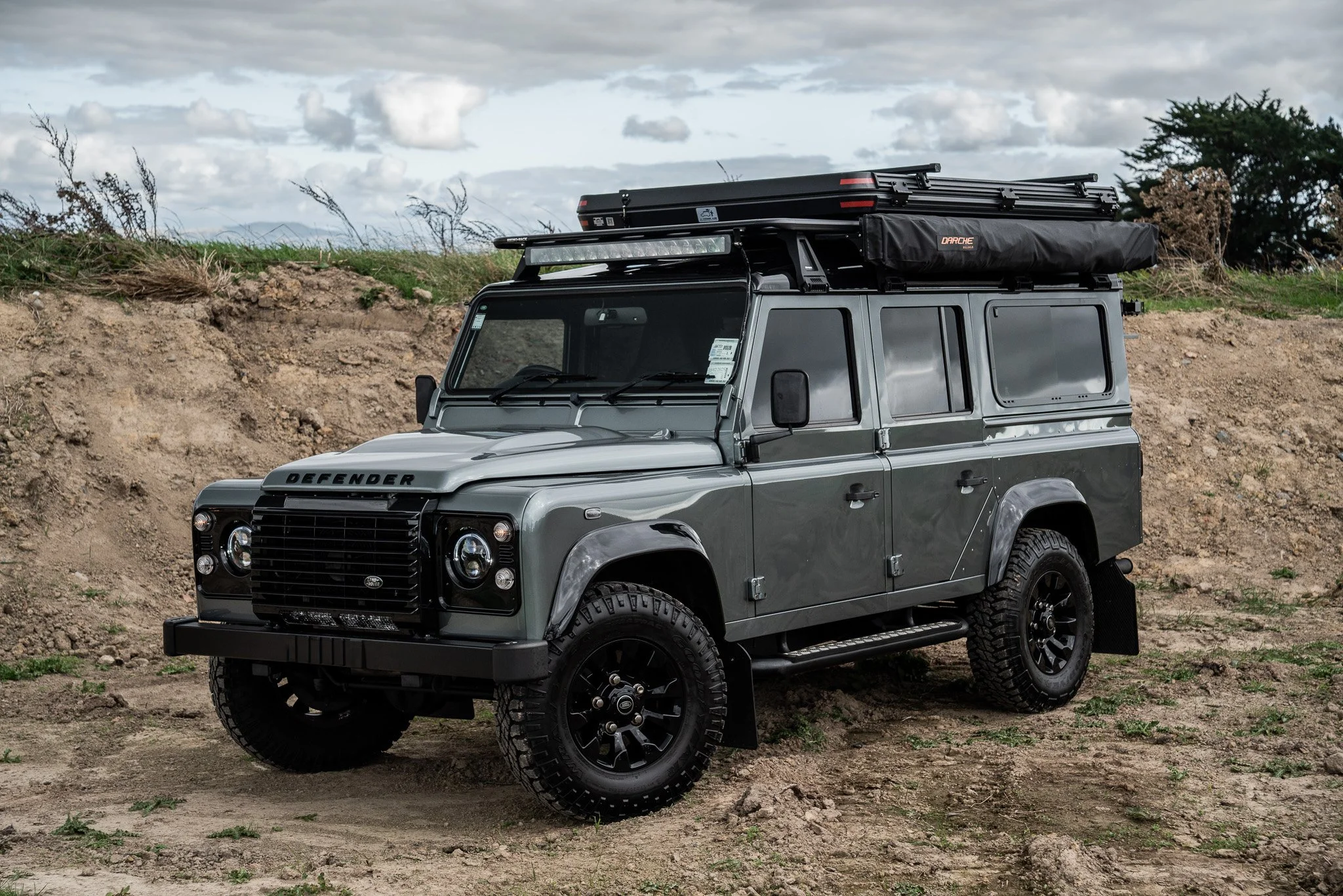 A gray Land Rover Defender parked on dirt ground outside with a cloudy sky in the background. Roof rack with camping gear and accessories.