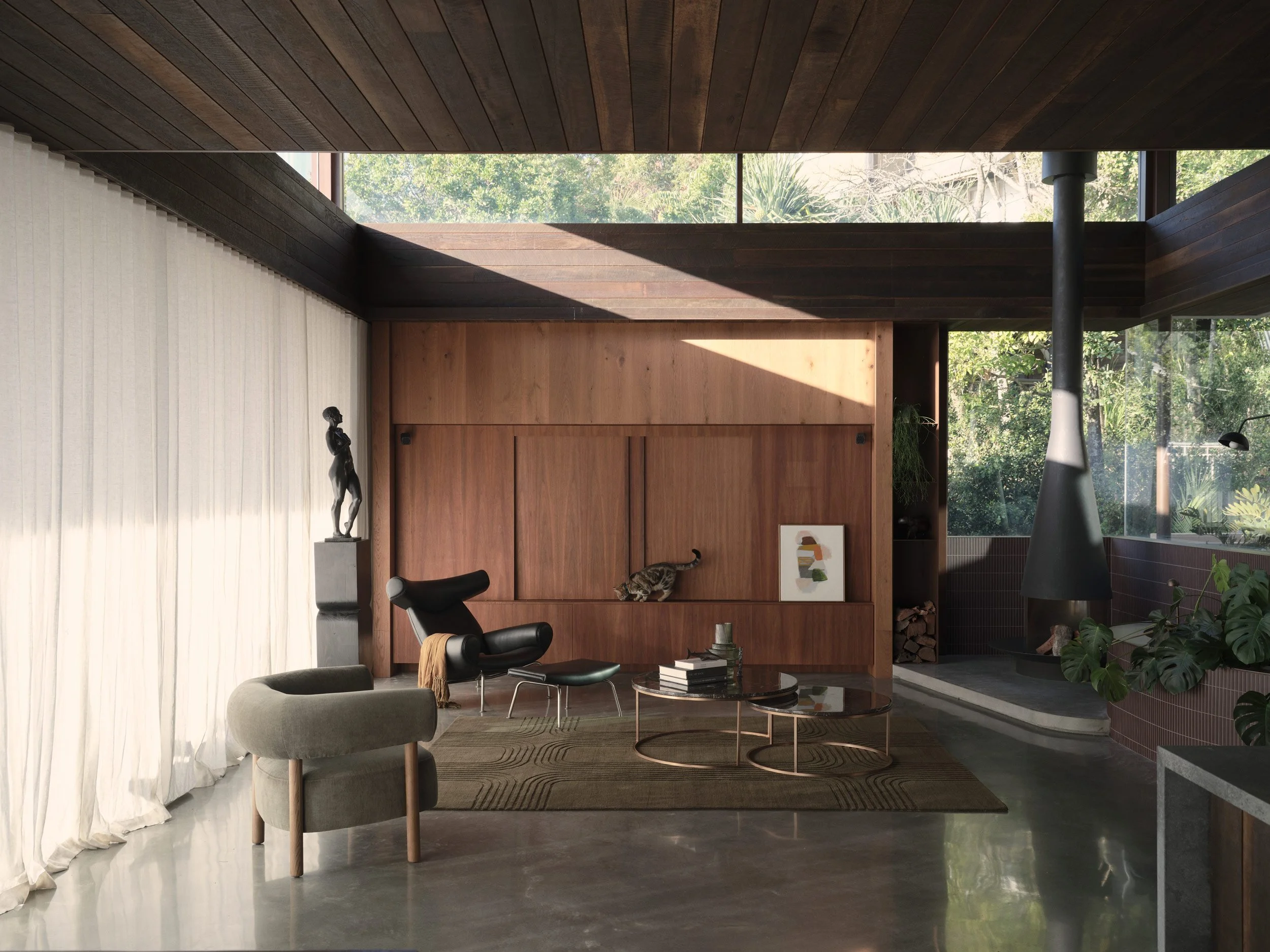 Modern living room with wooden wall panels, a black lounge chair, a beige armchair, a glass coffee table, a sculpture of a woman on a pedestal, and large windows showing greenery outside.