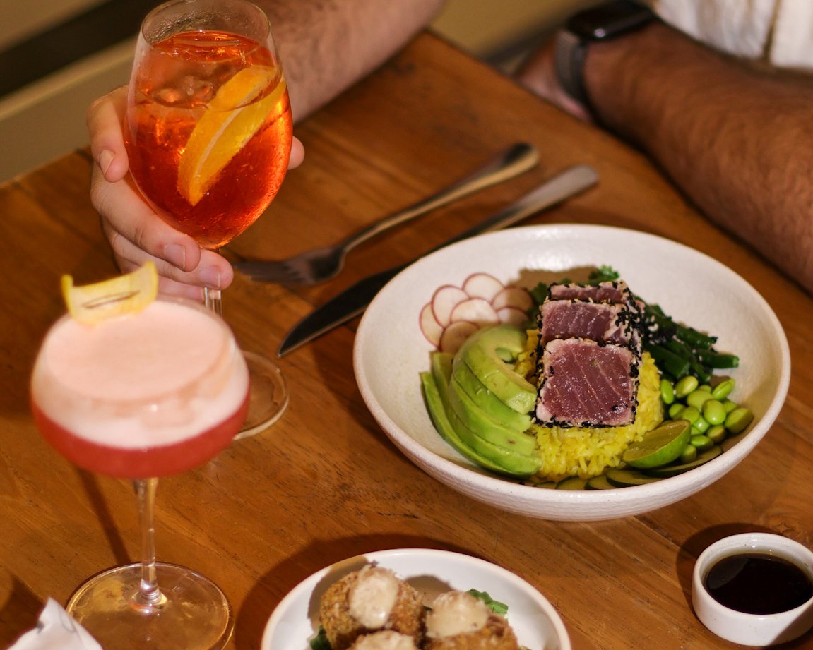 A person holding a glass of Aperol Spritz with an orange wedge, a colorful poke bowl with tuna, avocado, radishes, edamame, and rice, a plate of fried appetizers, and a small container of soy sauce on a wooden table.