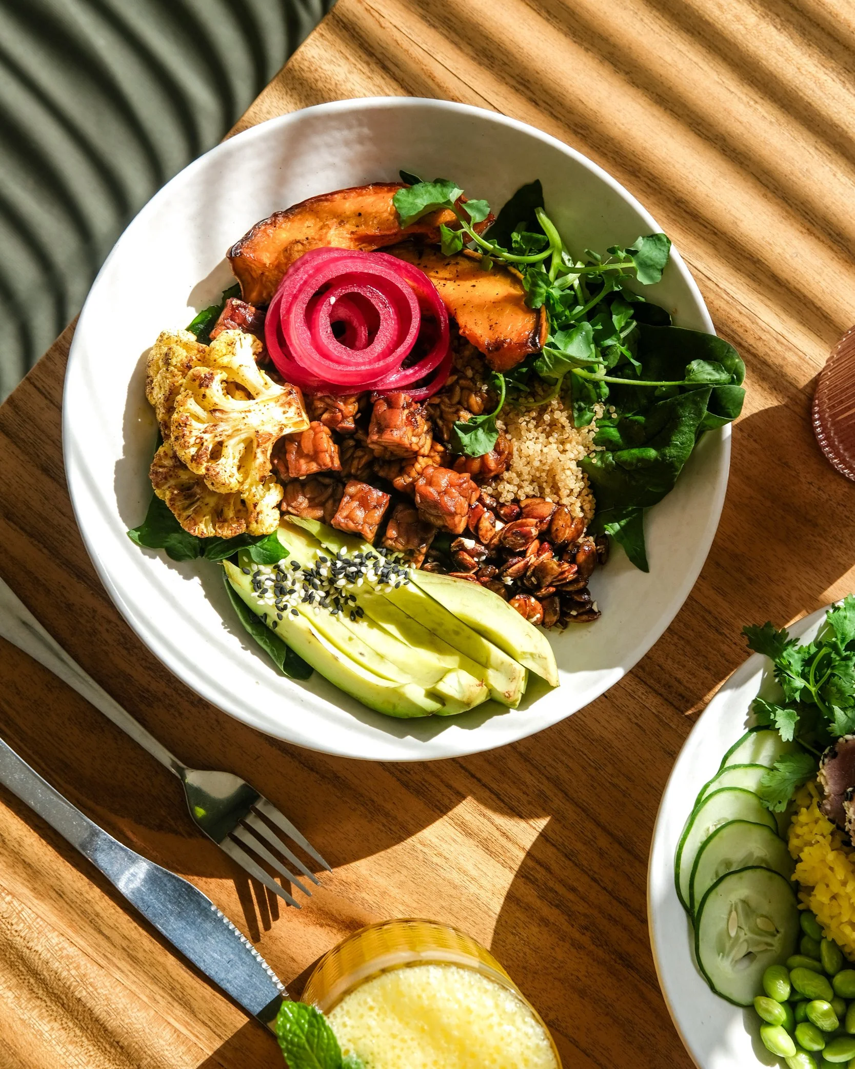 A colorful bowl of assorted healthy foods including grilled pumpkin, pickled onions, greens, cooked grains, roasted veggies, avocado slices with sesame seeds, and some nuts or seeds.