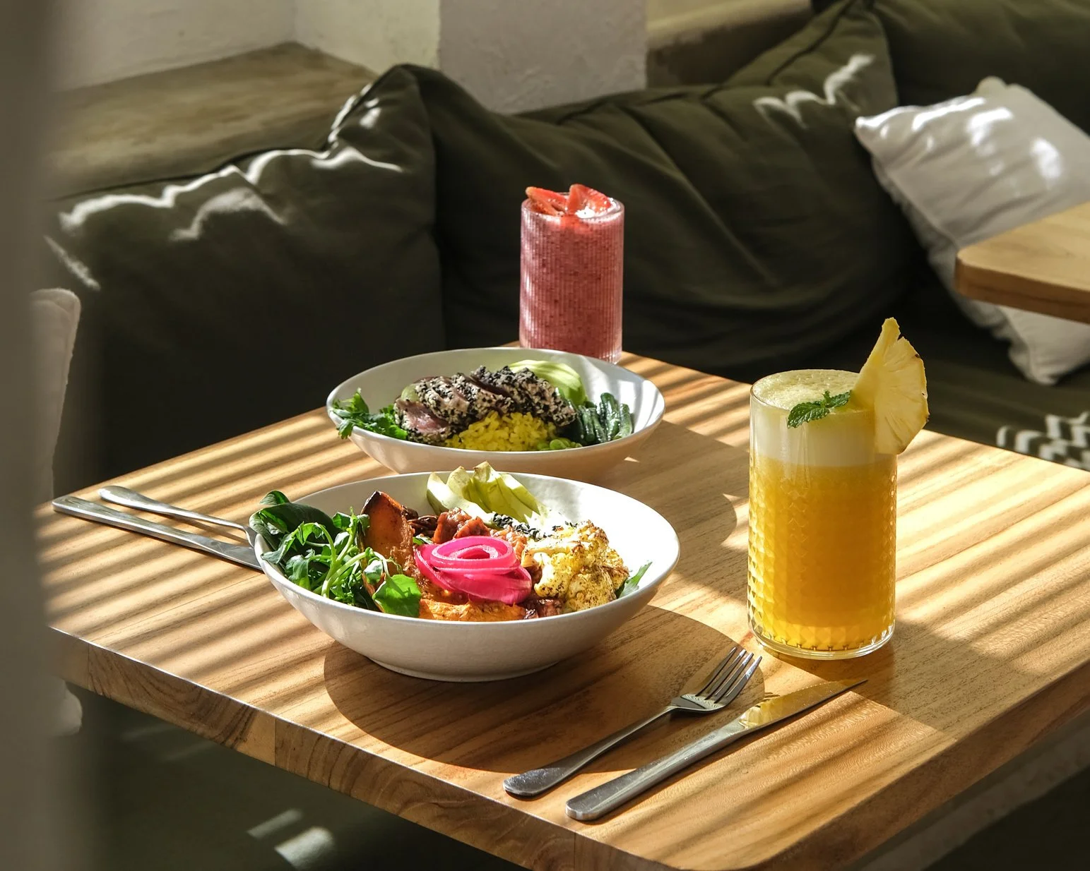 A wooden table with two bowls of food and two colorful drinks, sunlight casting shadows. The background shows a sofa with pillows.