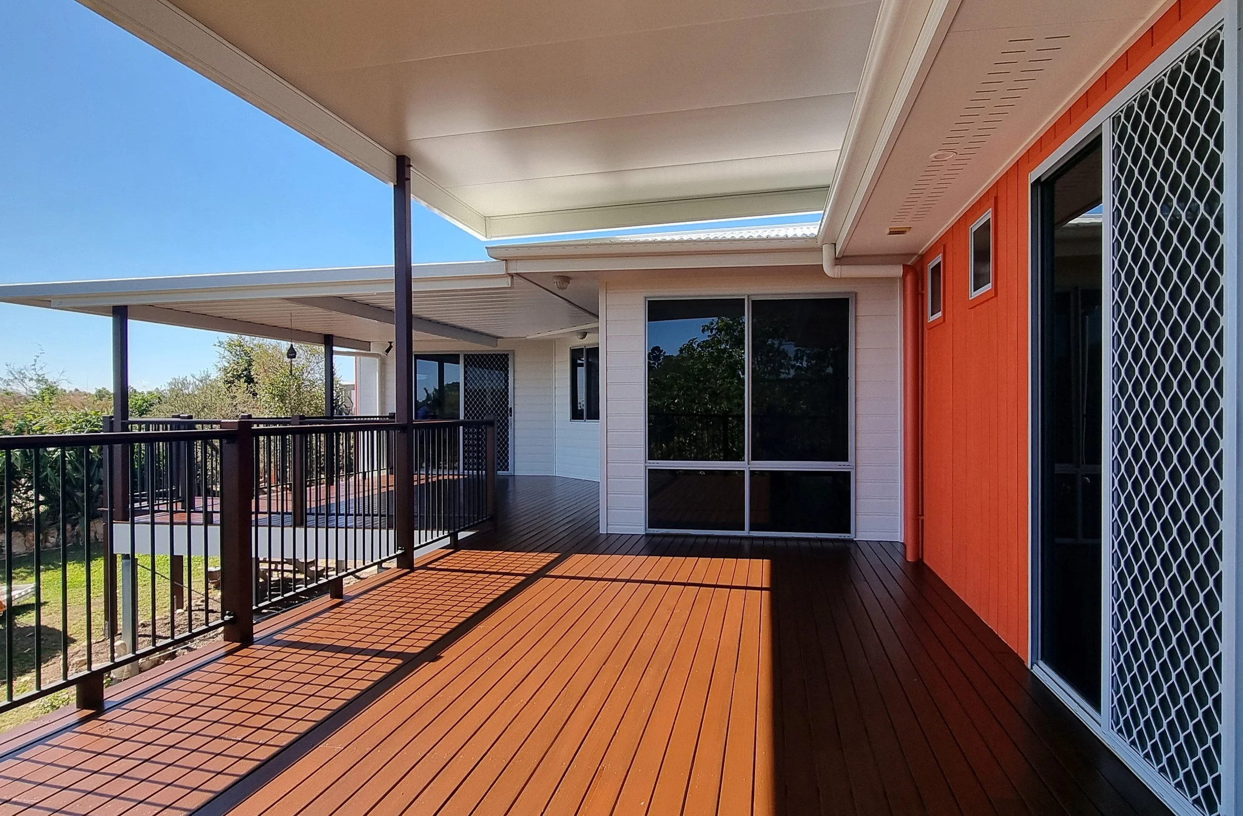 Outdoor balcony with wooden flooring, black railing, and a view of trees and sky.