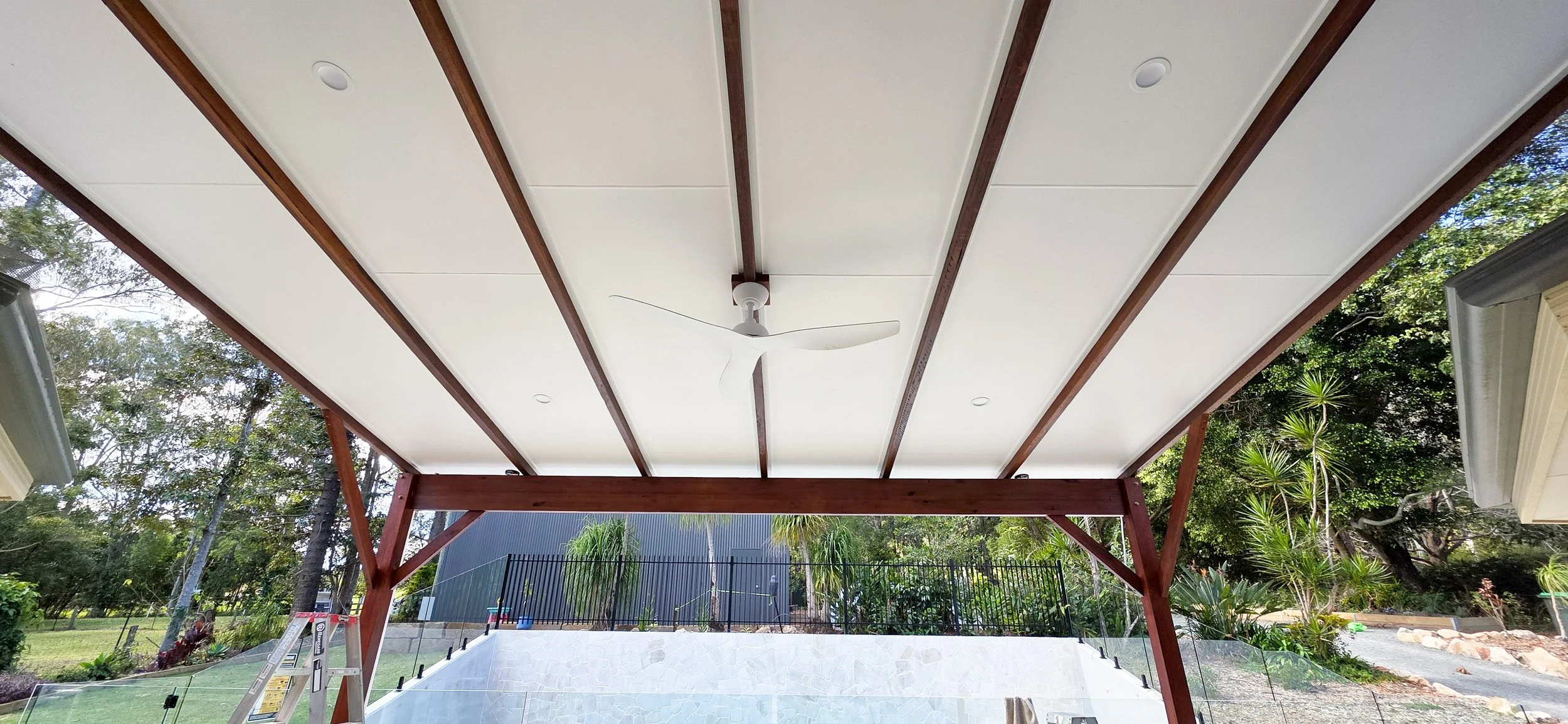 View of a covered outdoor patio with white ceiling, wooden beams, a ceiling fan, and outdoor greenery in the background.