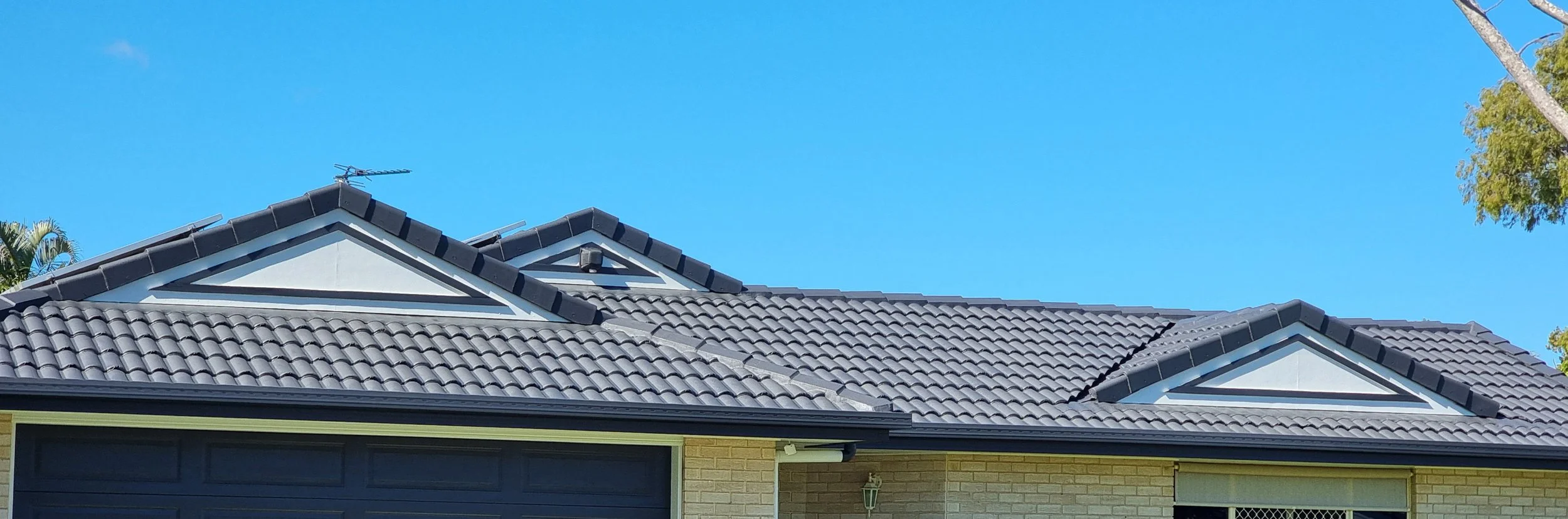 Close-up view of a house roof with dark gray tiles, triangular dormer windows with white trim, and a clear blue sky in the background.