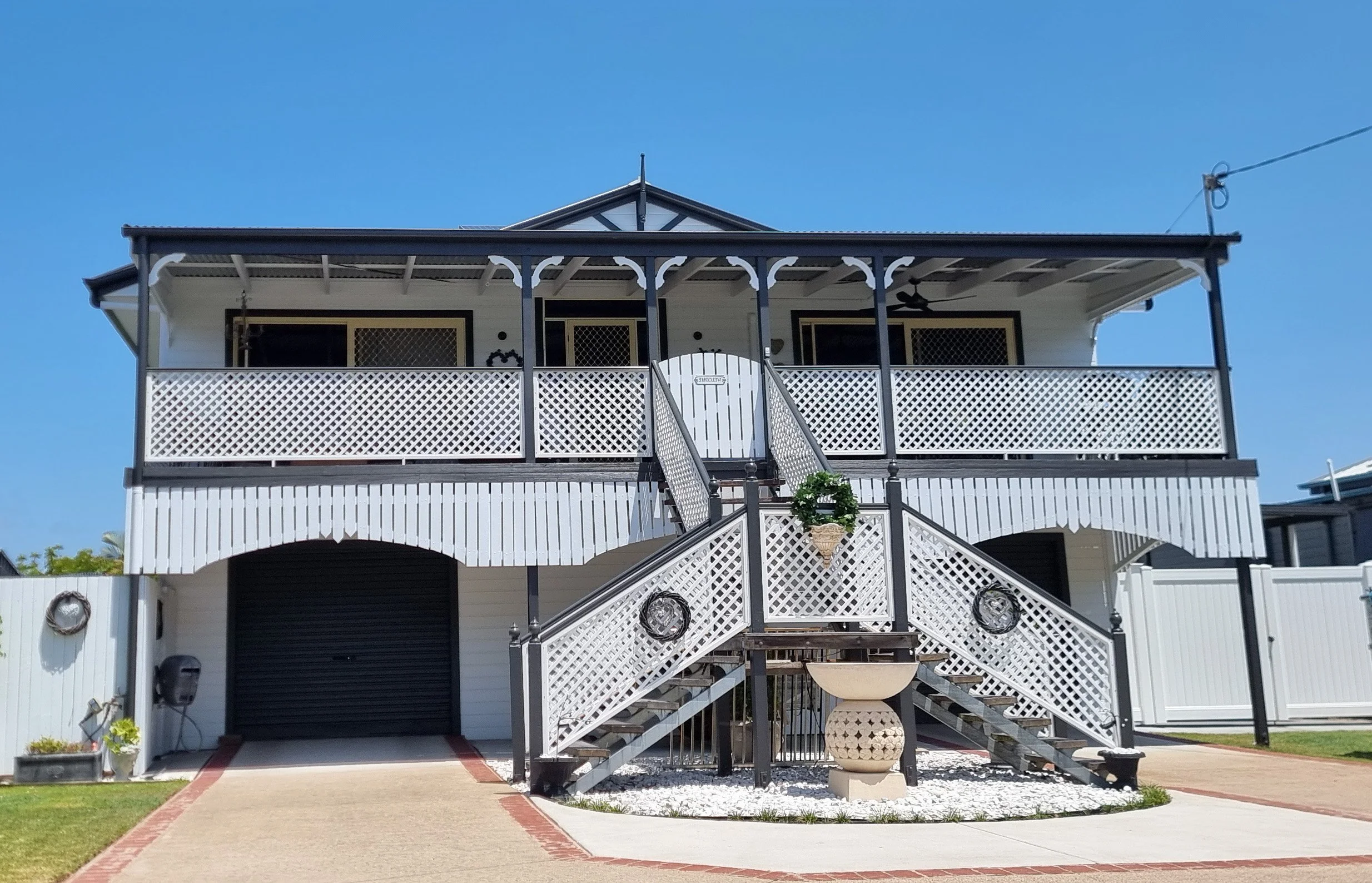 Two-story house with a white exterior and black trim, a staircase leading to the upper deck with white lattice railings, and a garage on the lower level. Clear blue sky in the background.