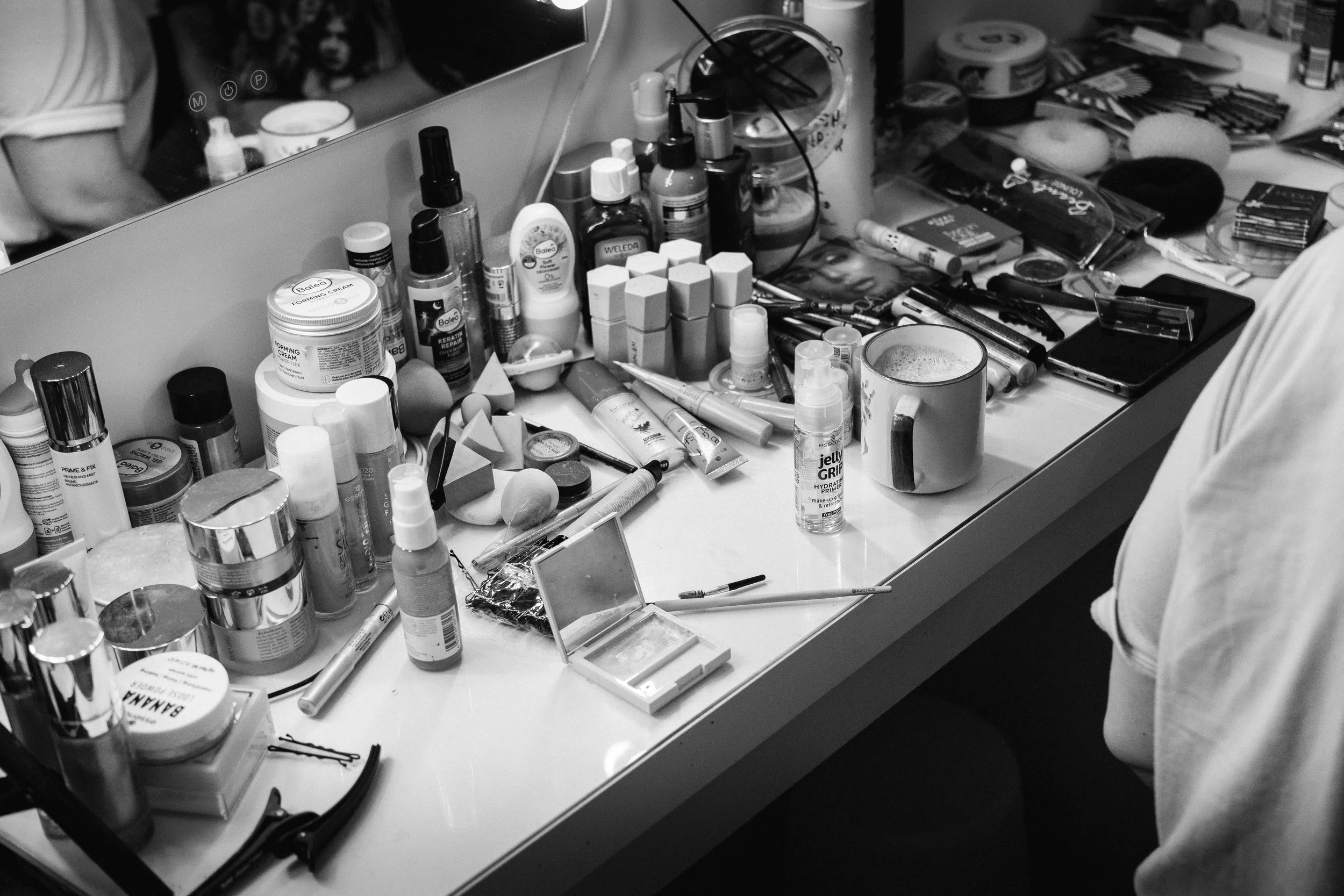 Messy makeup vanity table with various cosmetics, brushes, skincare products, and a mirror, with part of a person in a light-colored shirt visible on the right side.