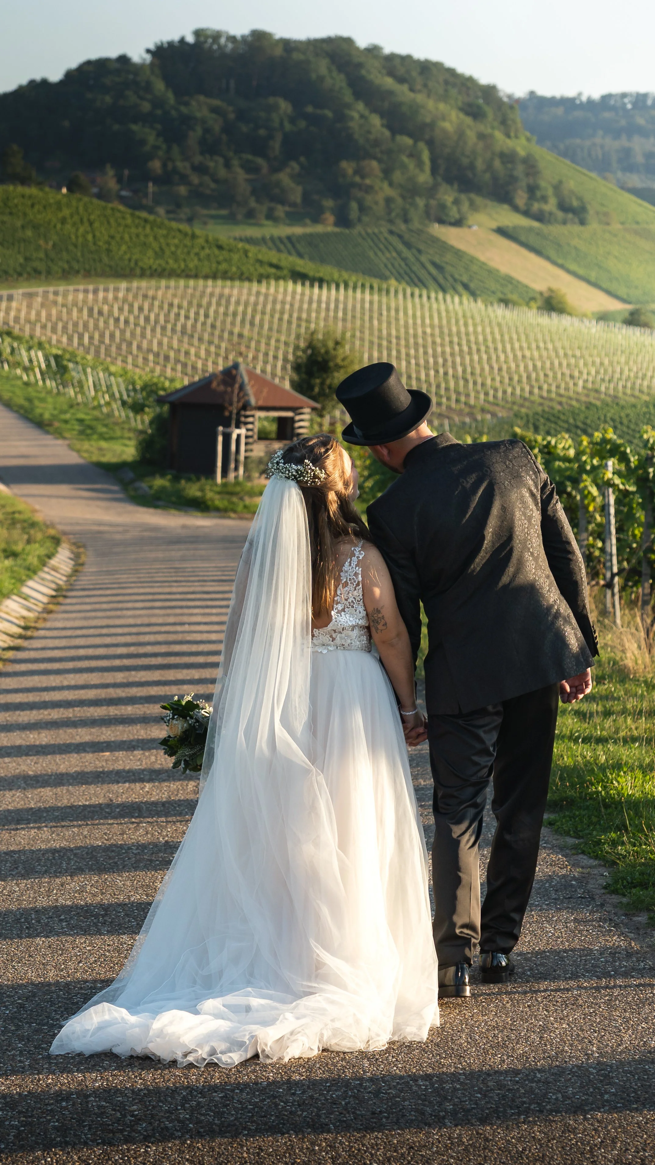 A newlywed couple holding hands and walking on a vineyard road with lush green hills in the background during sunset.