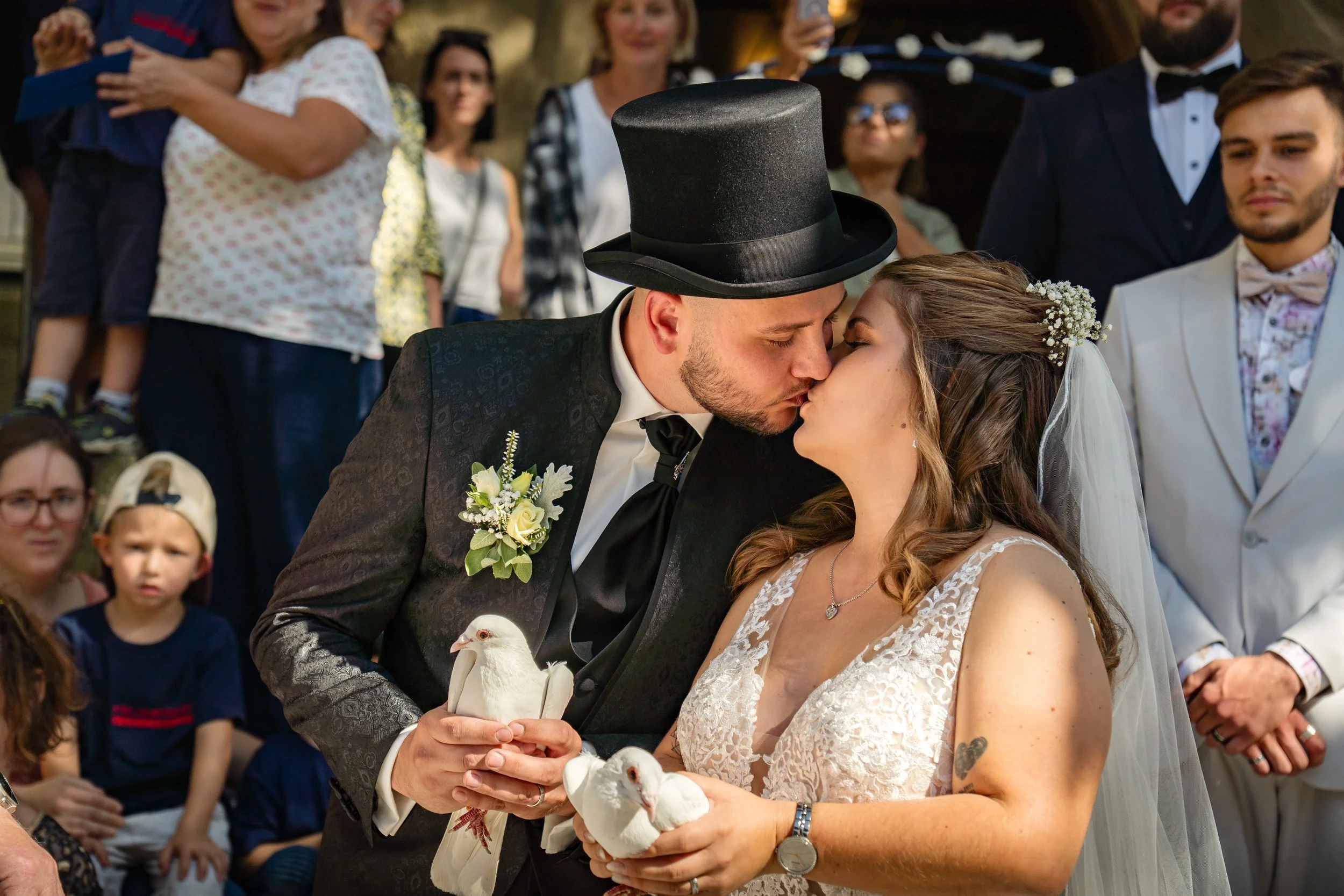 A bride and groom wearing a top hat share a kiss during their wedding ceremony, holding white doves in their hands, with wedding guests gathered around.