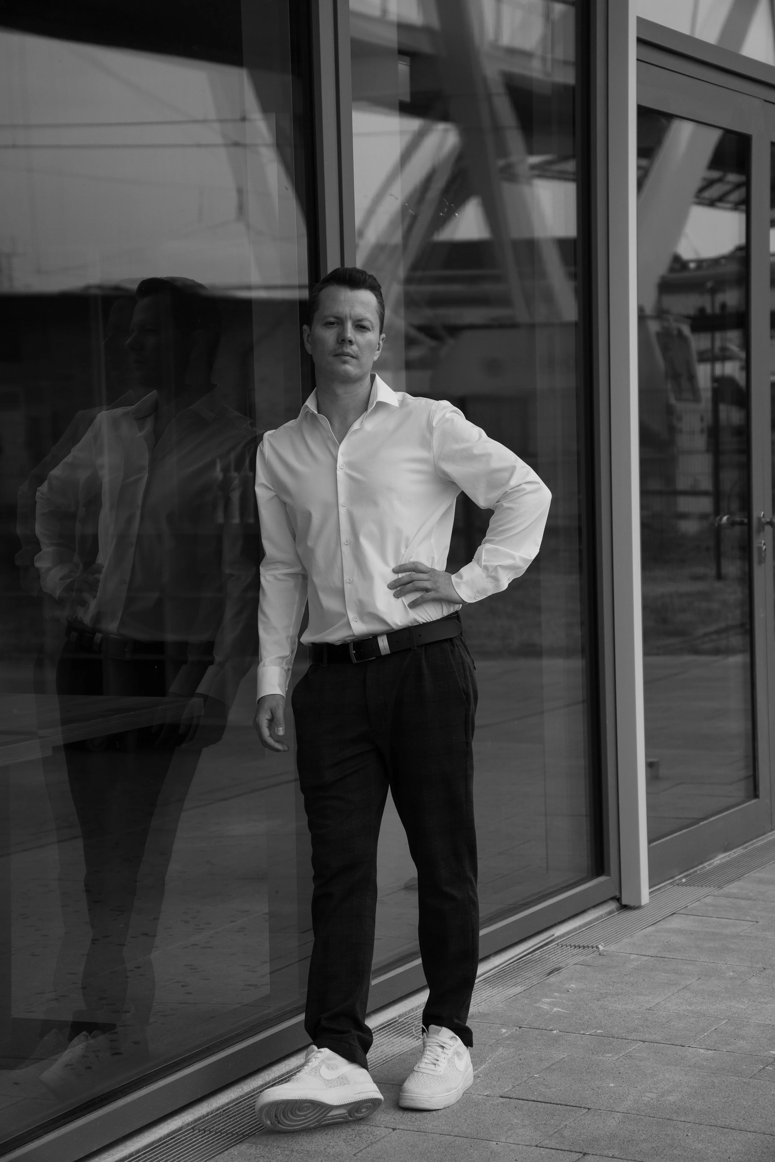 A man in a white shirt and dark pants standing outside a glass building, leaning against the glass with a hand on his hip, in black and white.