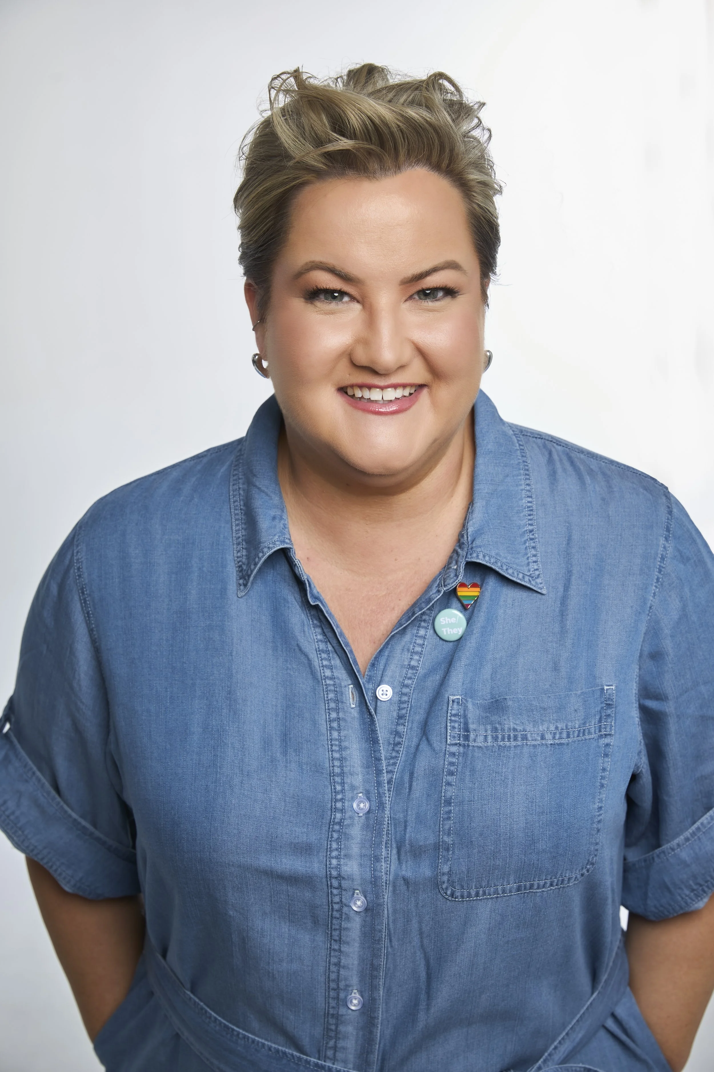 Mel Wyllie, a nonbinary femme-presenting person with short, styled blonde hair wearing a blue denim shirt, smiling at the camera, against a plain light background. She has a rainbow pride pin, a button with she/her pronouns