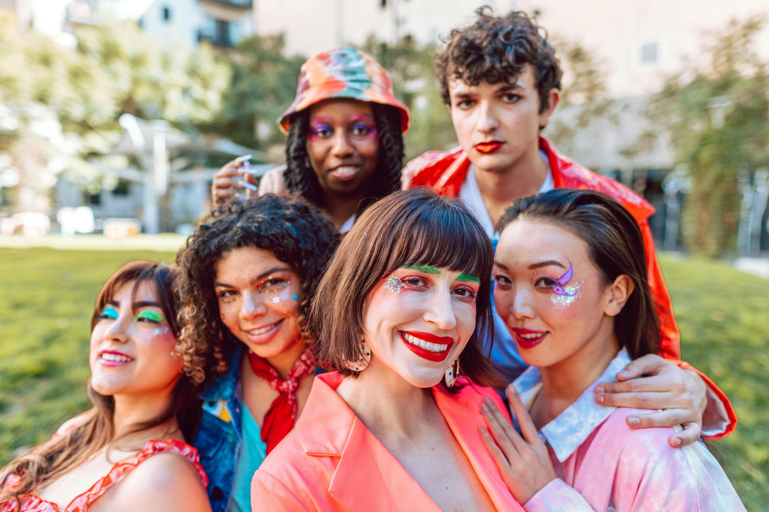 Group of friends in colorful outfits with neon makeup, smiling at the camera in a park.