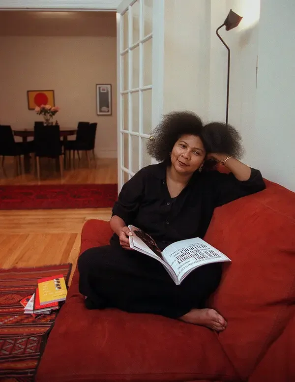 bell hooks sitting on a red sofa reading a magazine in her living room with a dining area in the background.