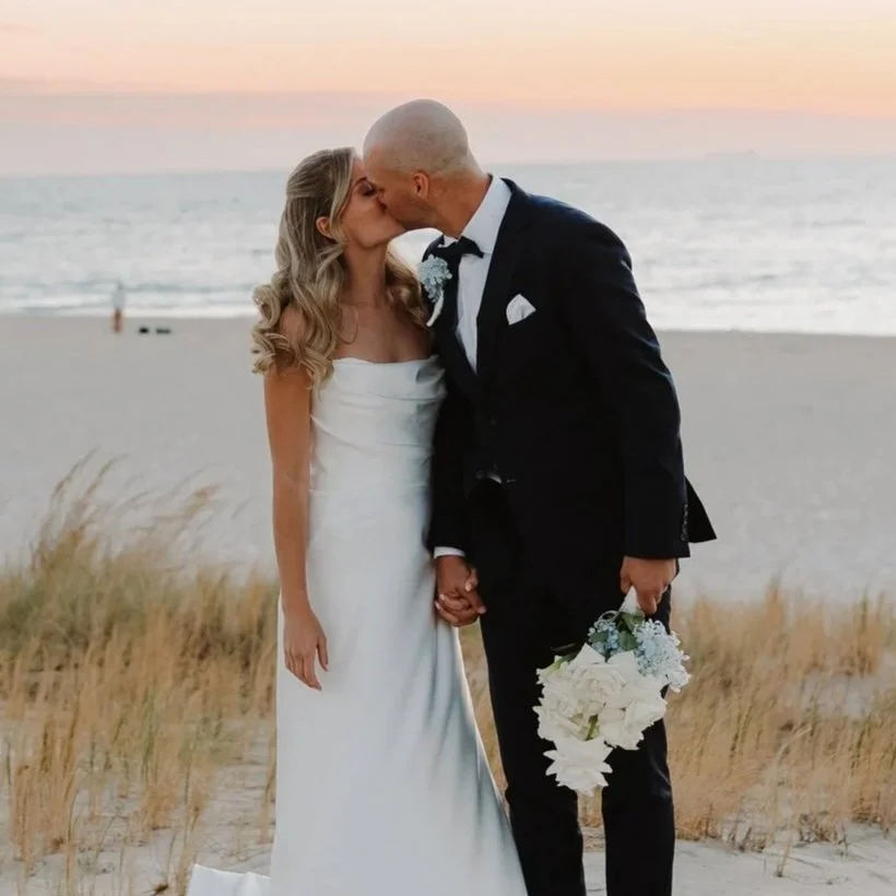 A newlywed couple kissing on a beach at sunset, the woman in a white wedding dress, the man in a black tuxedo holding a bouquet.