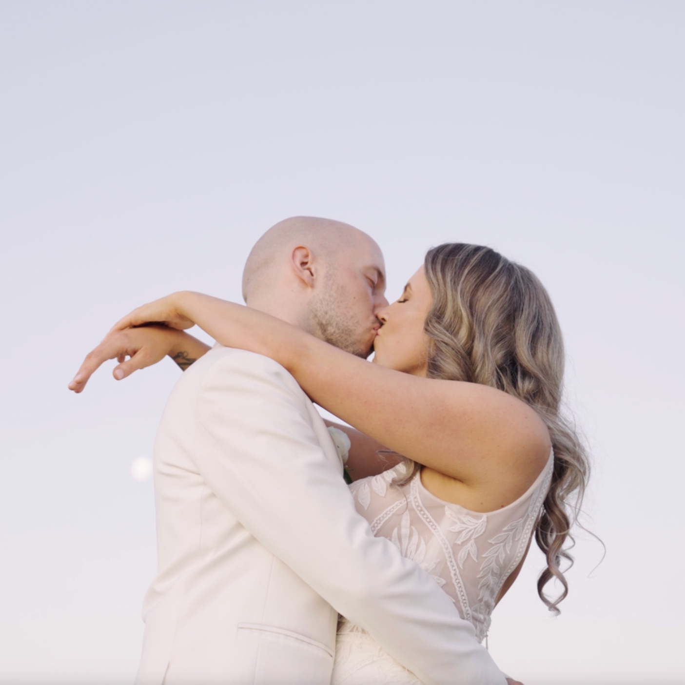 A couple kissing, with the woman wearing a lace wedding dress and the man in a white suit, against a plain sky background.