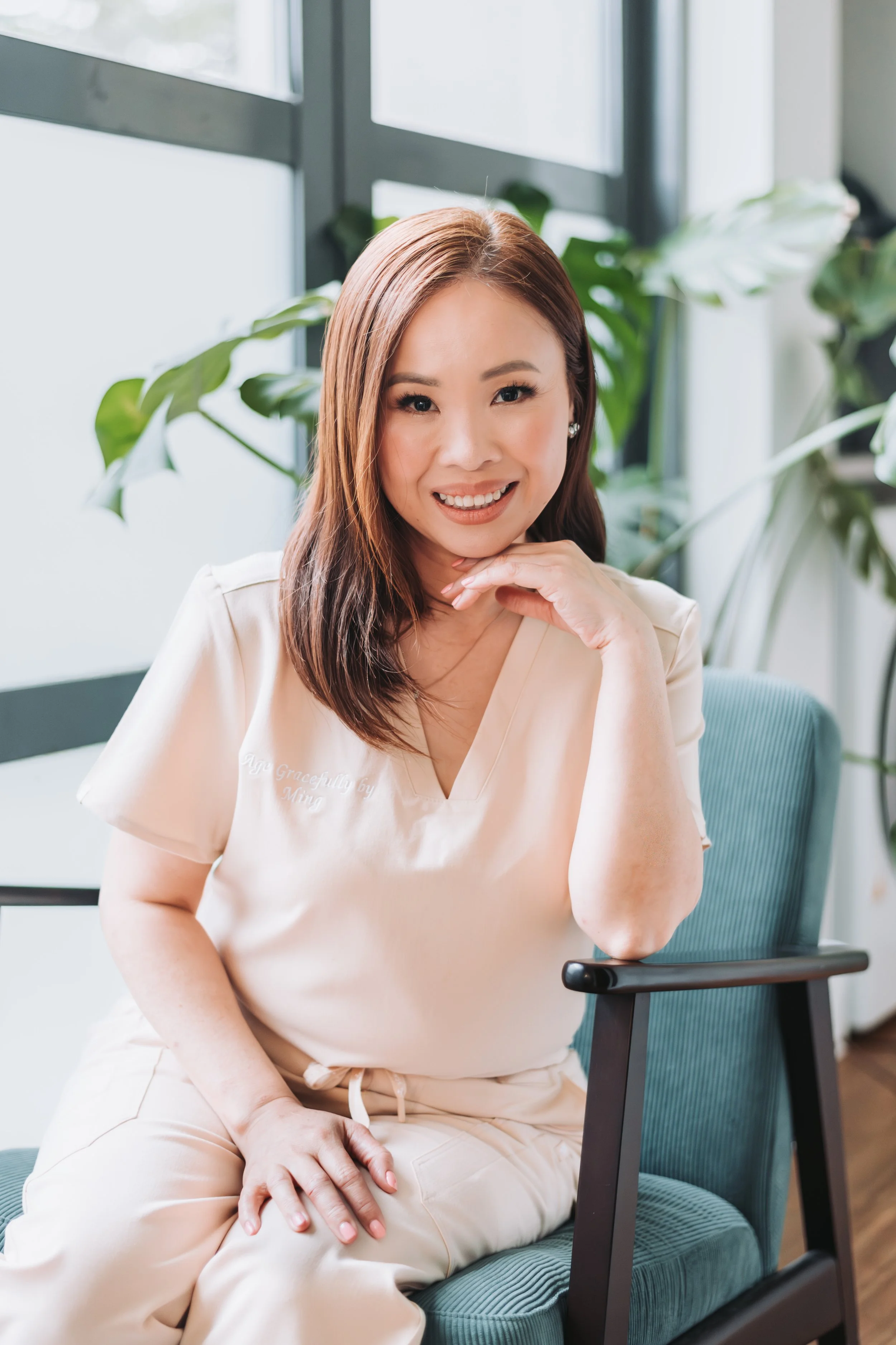 A woman with brown hair and light skin smiling while sitting on a teal chair with her chin resting on her hand, in an indoor setting with large windows and green plants in the background.
