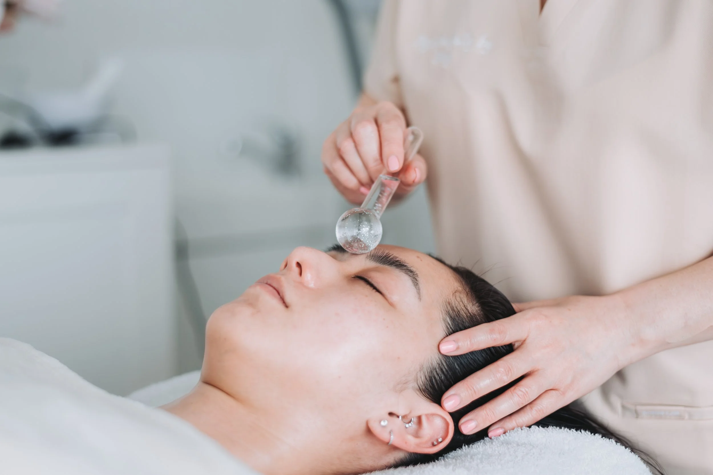 A woman receiving a facial treatment with a glass cooling globe, applied to her forehead, while lying down with her eyes closed.