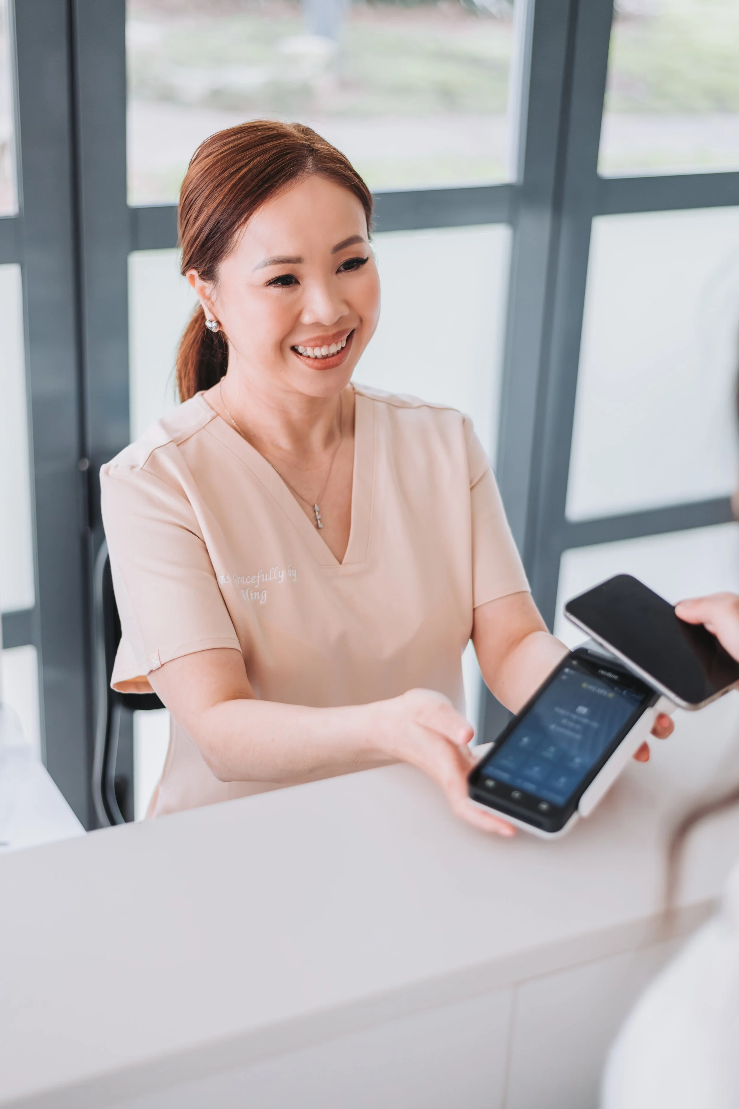 A woman in beige medical scrubs smiling and handing a smartphone to a person across a reception desk in a medical or professional setting.