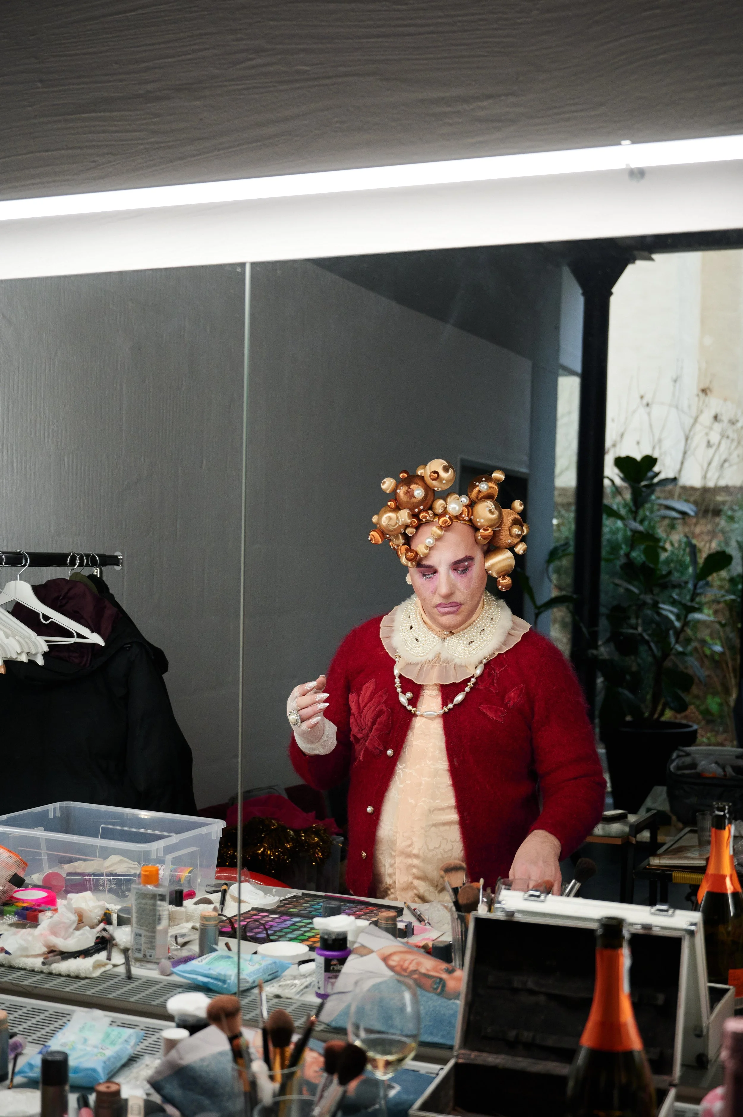 A person dressing in elaborate makeup and costume, with a headpiece made of golden and pearl-like ornaments, standing in front of a mirror surrounded by makeup products and brushes.