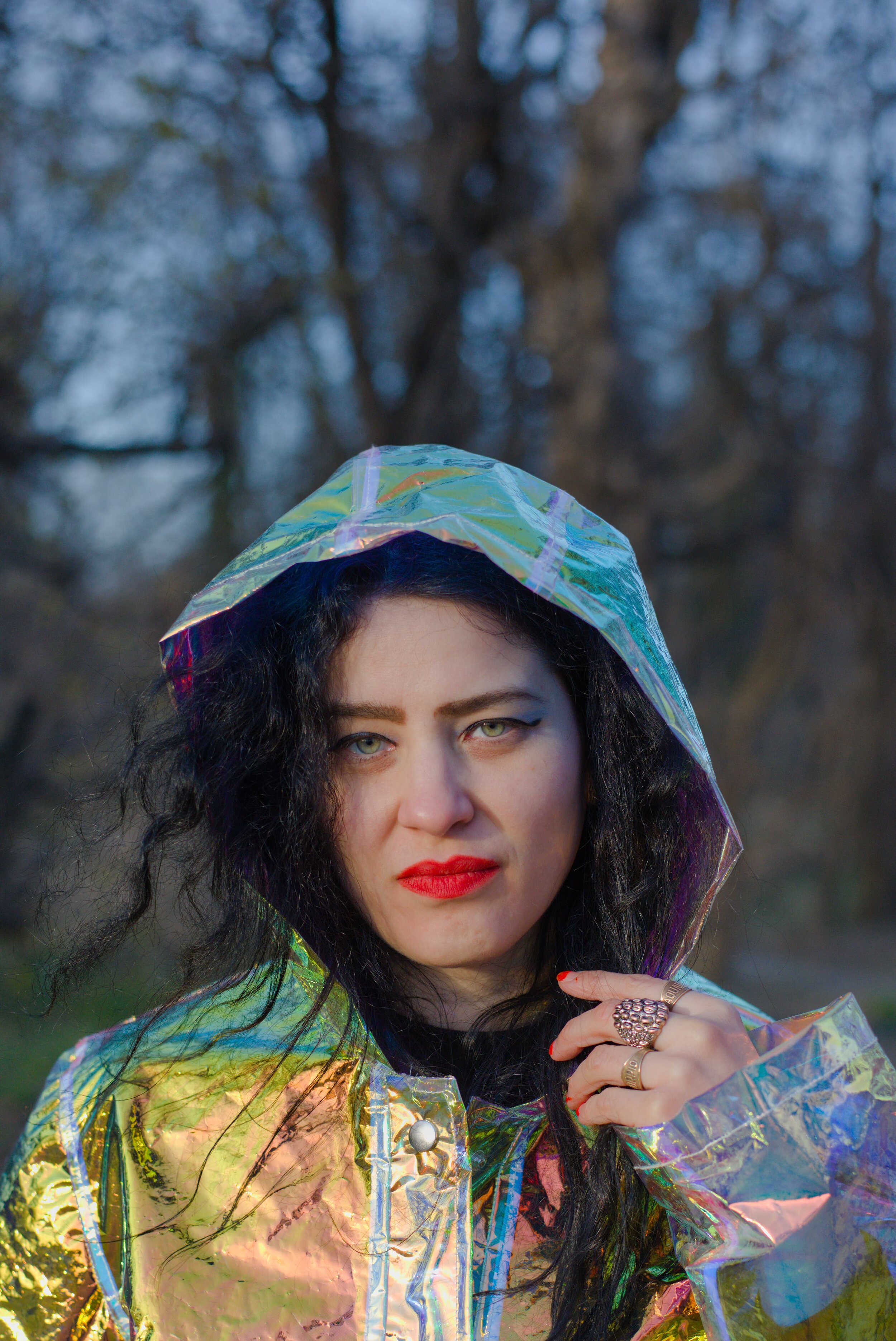 A woman with black, curly hair and striking makeup, wearing a shiny, iridescent raincoat with the hood up, standing outdoors with blurred trees in the background.