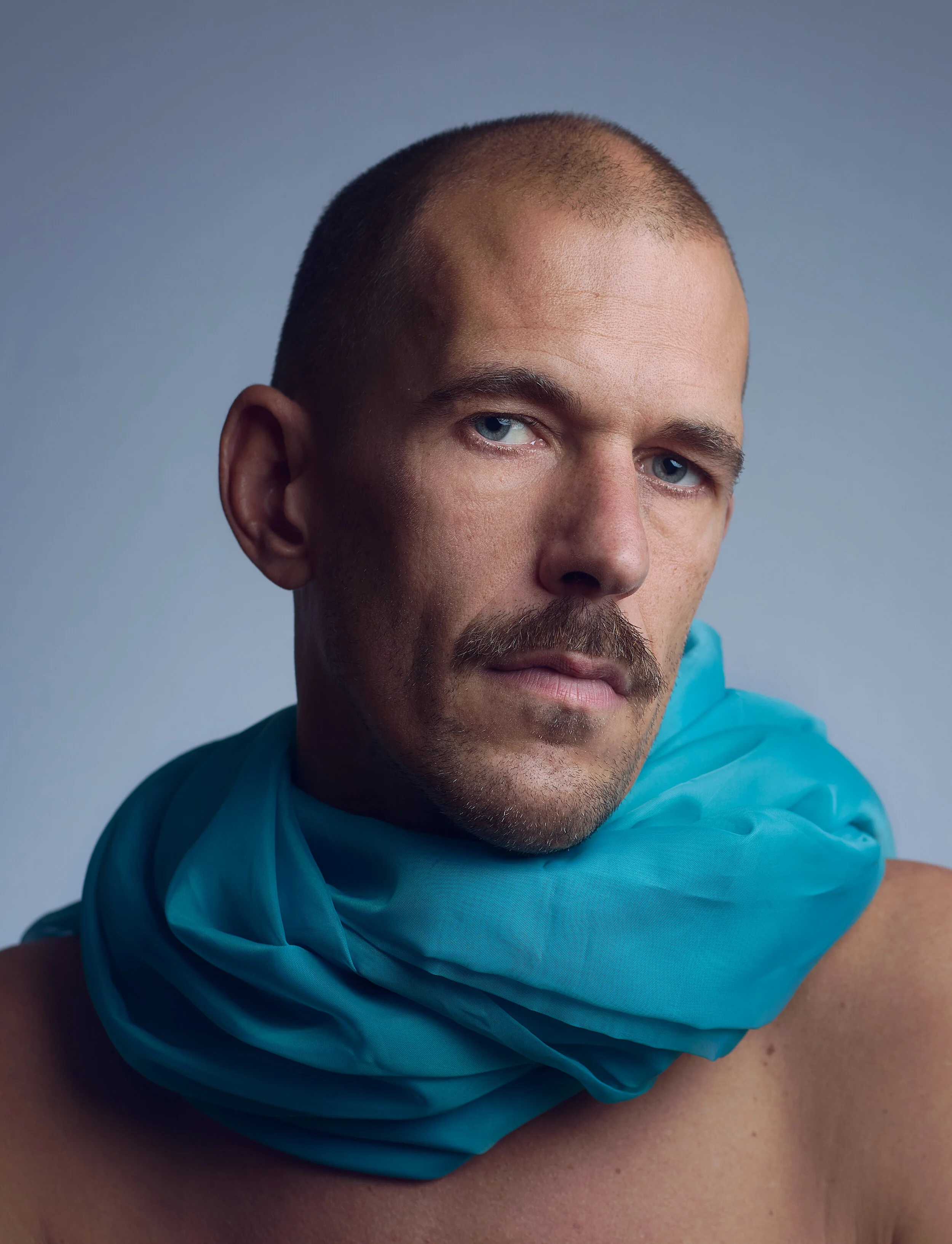 Close-up portrait of a man with blue eyes and a mustache, wearing a blue scarf, with a neutral background.