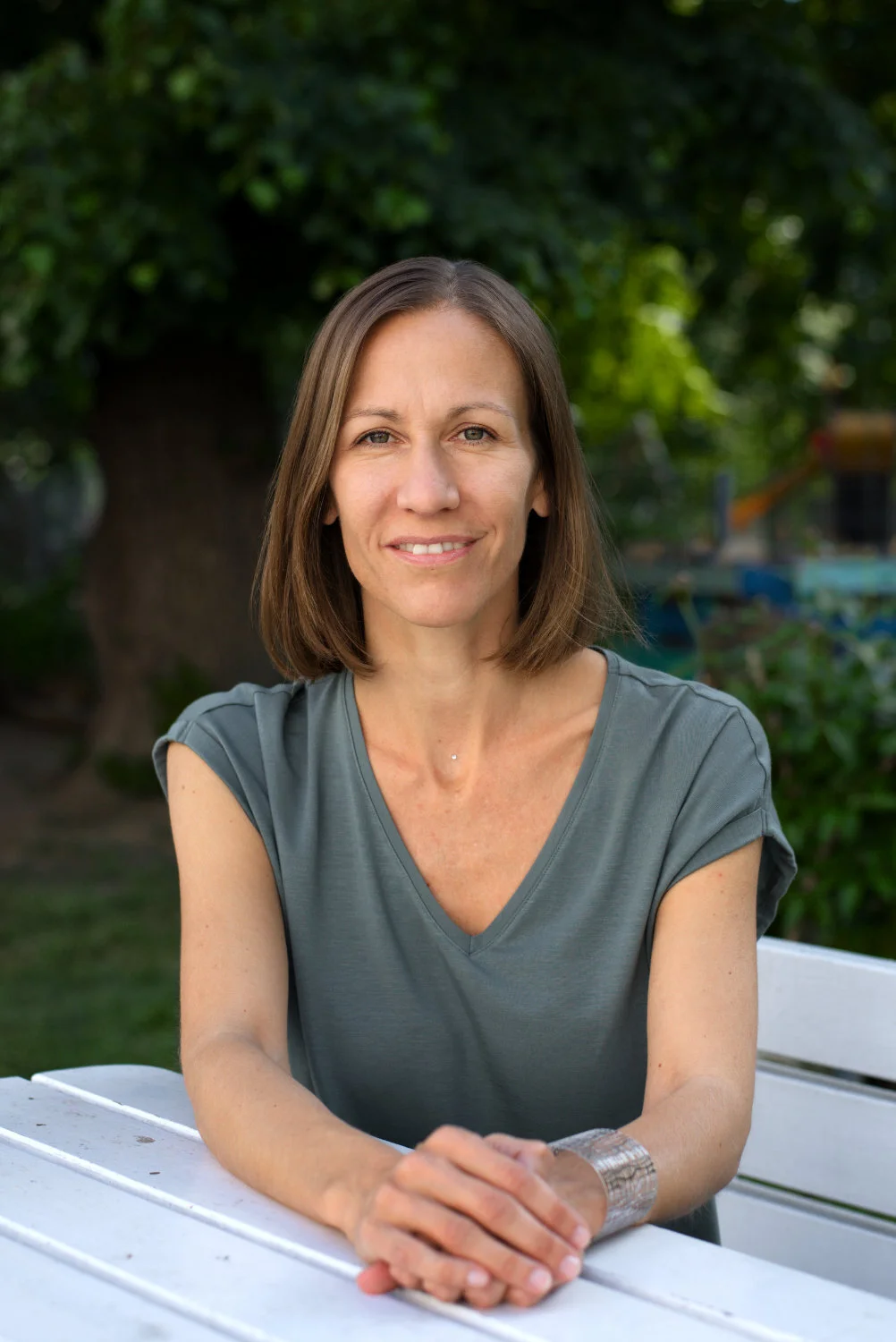 Woman with brown hair smiling, sitting outdoors at a white table, greenery in the background.