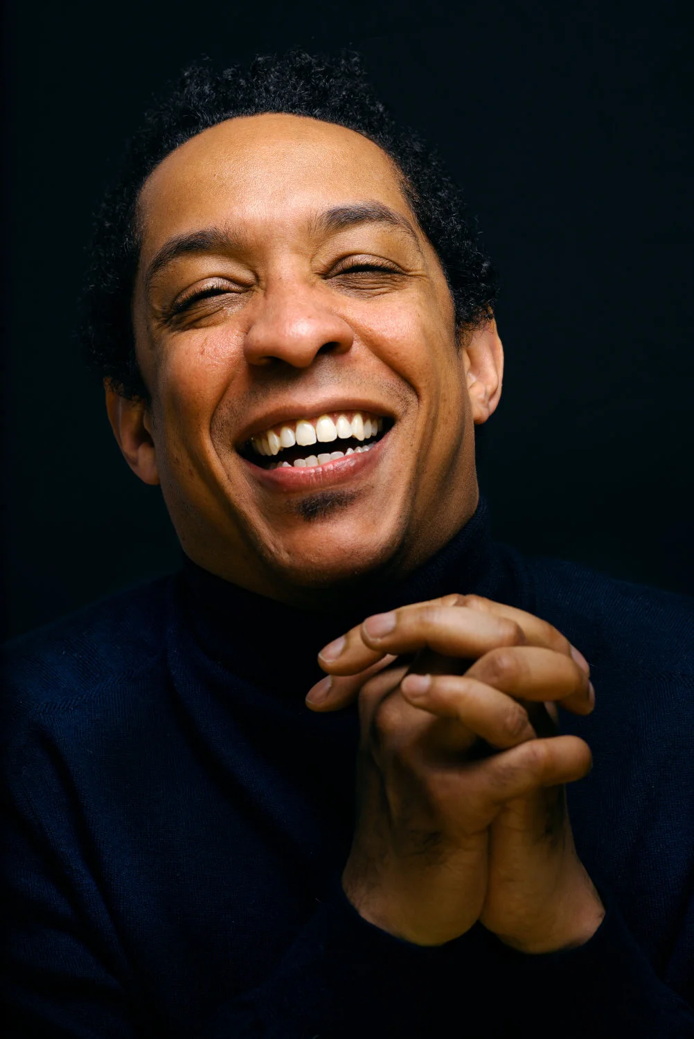 A close-up portrait of a smiling man with curly hair, clasping his hands, wearing a dark turtleneck against a black background.