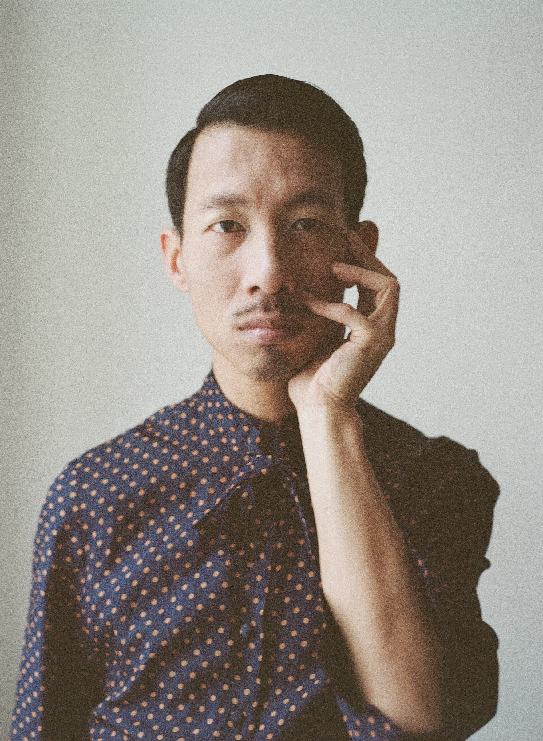 A man with short black hair and a slight mustache, wearing a navy blue shirt with orange polka dots, is touching his face with his right hand, looking directly at the camera against a plain light background.