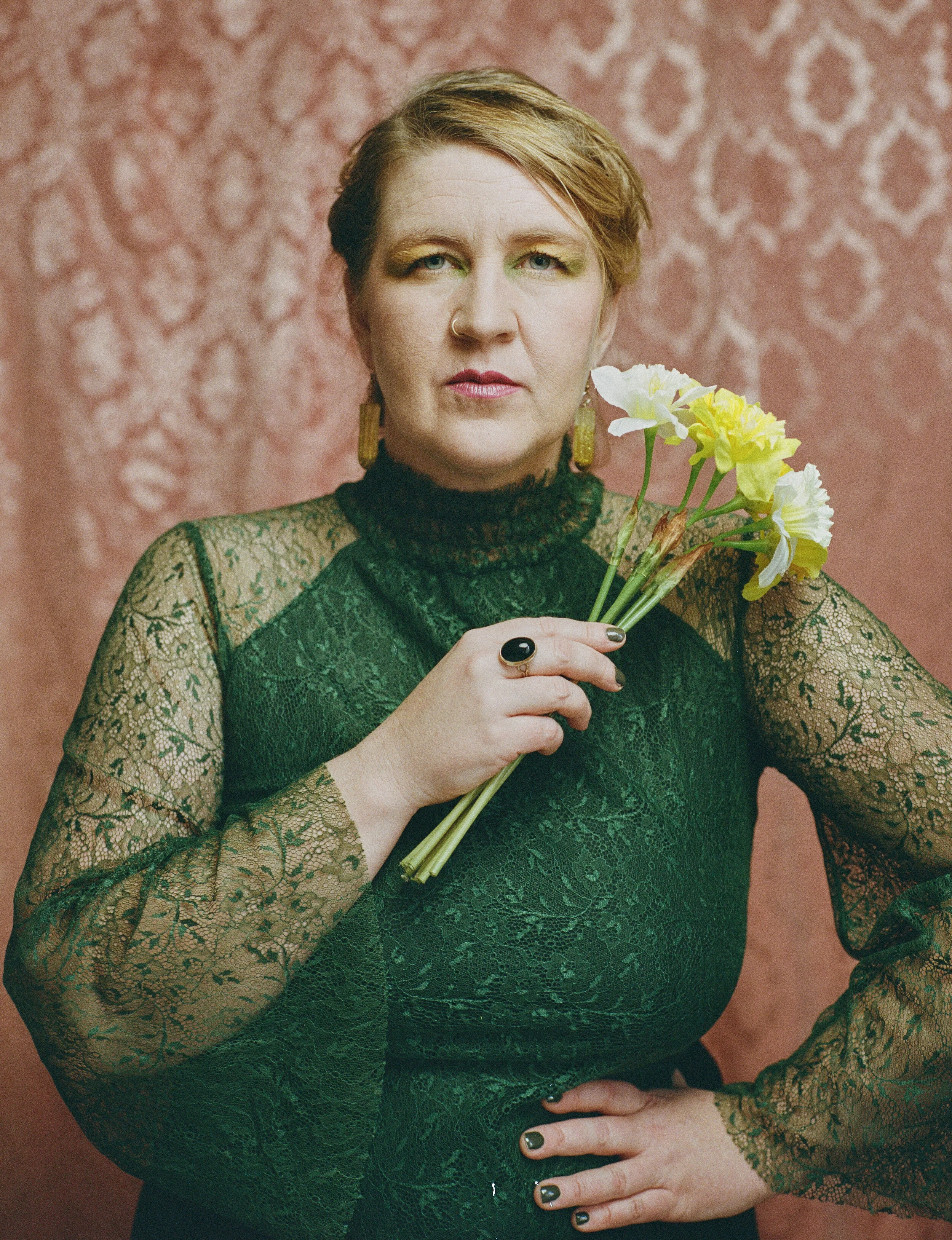 A woman with short, styled hair and light skin holding a small bouquet of yellow and white flowers against a pink patterned background. She is wearing a black lace top with long, sheer sleeves, gold earrings, a black ring, and has dark nail polish.