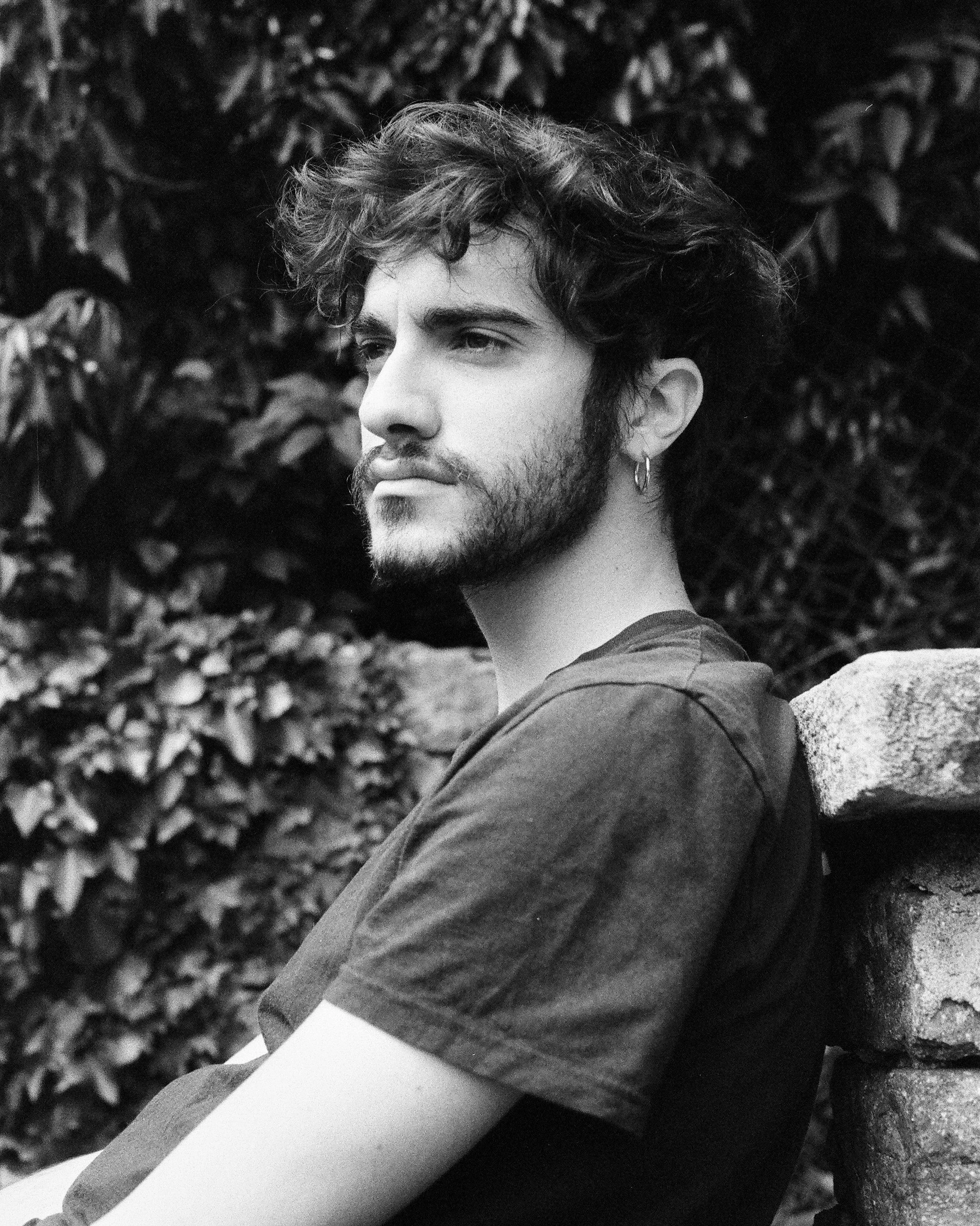 A young man with curly hair, a beard, and piercings, sitting outdoors next to a stone wall, looking contemplative, in black and white.