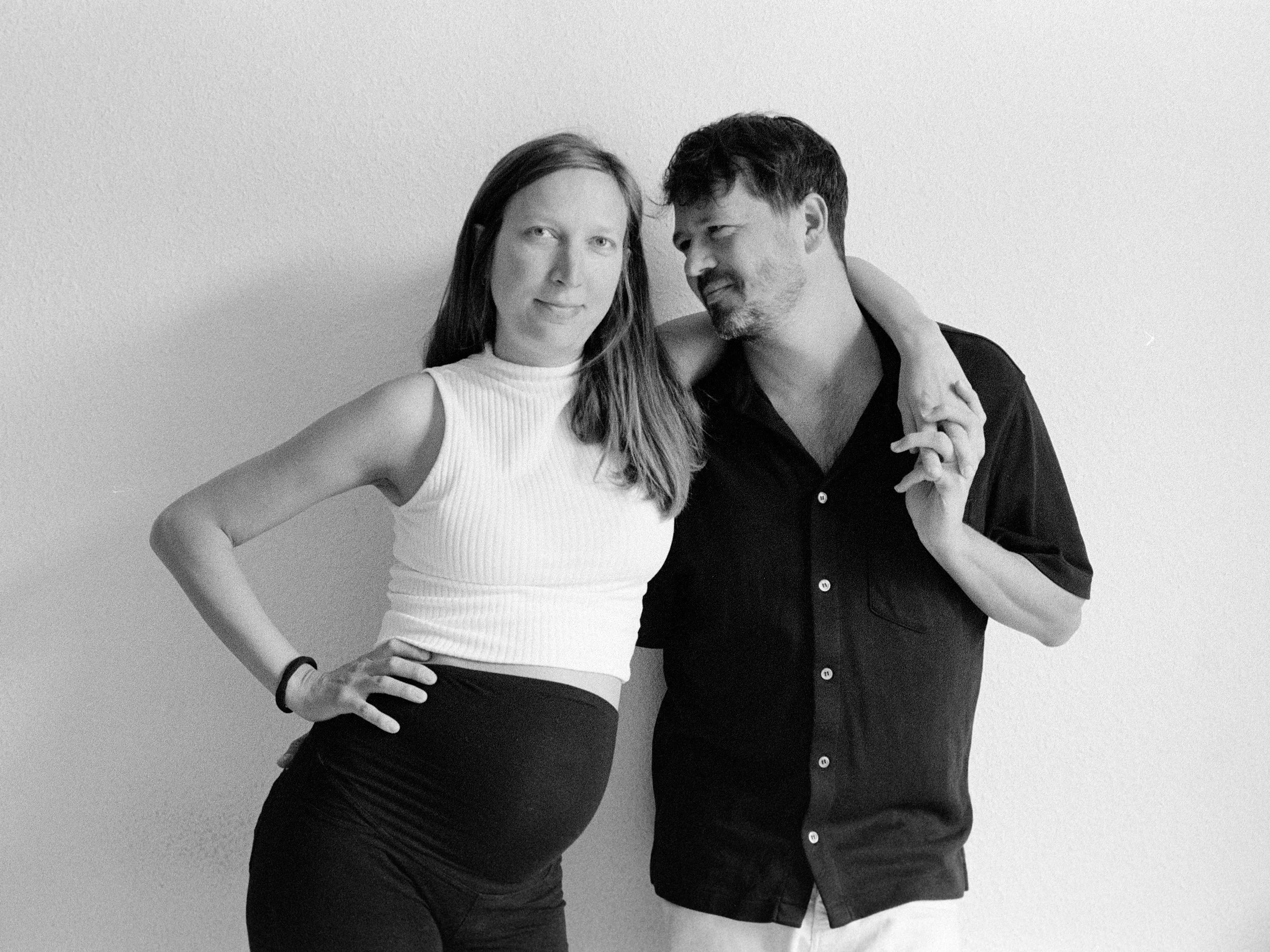 A black and white photo of a pregnant woman with long hair, wearing a sleeveless ribbed top and high-waisted pants, standing with her hand on her hip. A man with short hair and a beard, wearing a button-up shirt with rolled-up sleeves, stands beside 