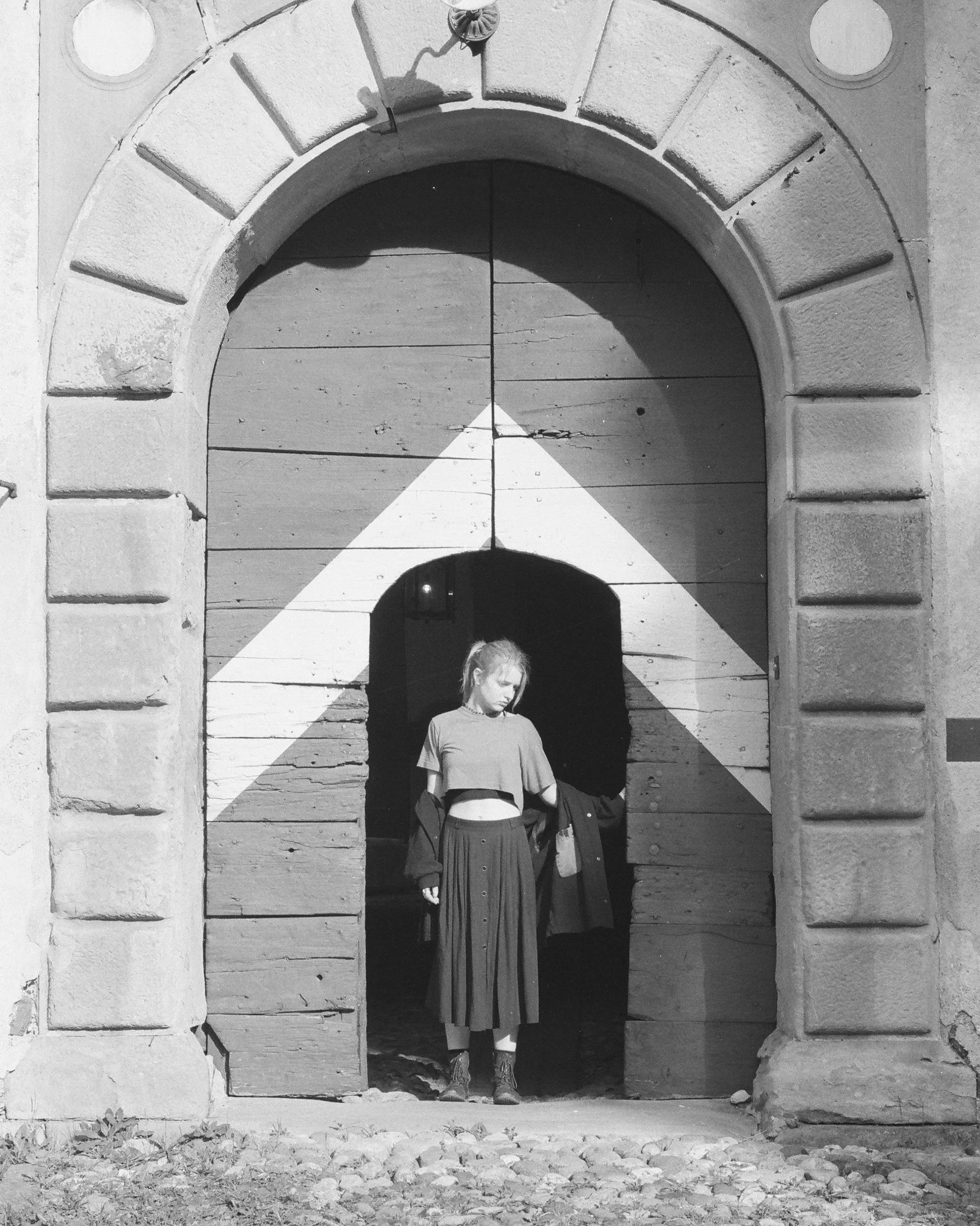 A young woman in a cropped T-shirt and long skirt standing in an arched doorway, with sunlight casting a triangle-shaped shadow on a wooden wall behind her.