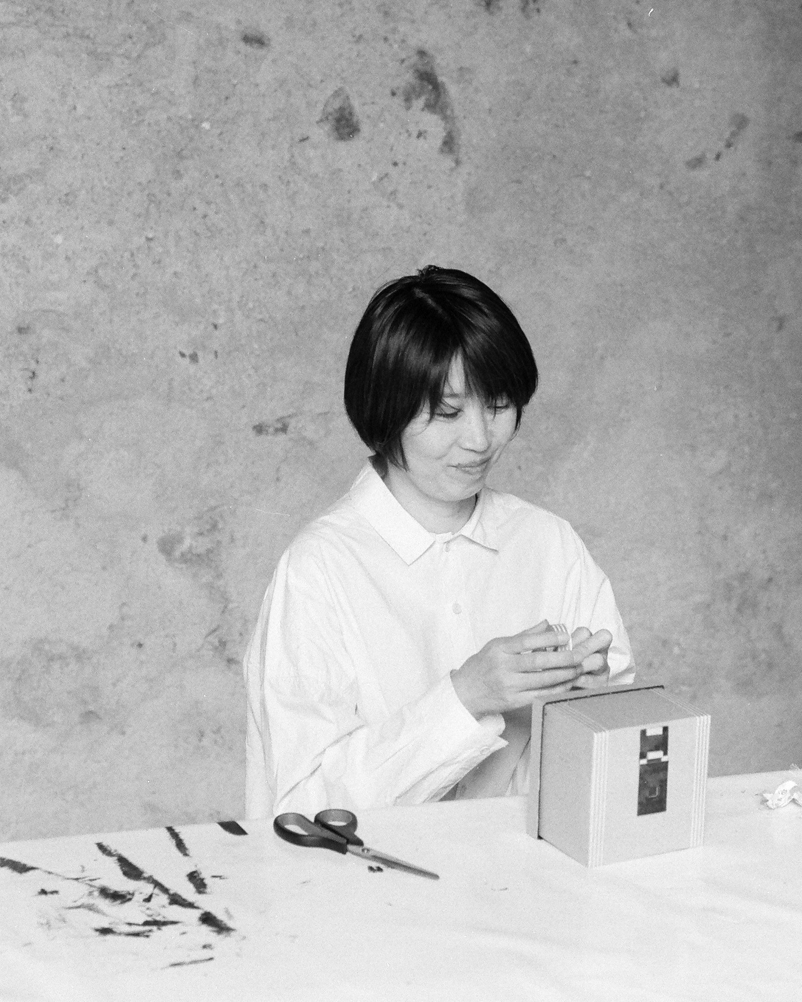 A woman sitting at a table, opening a box with scissors and ink on the table, against a textured wall background.