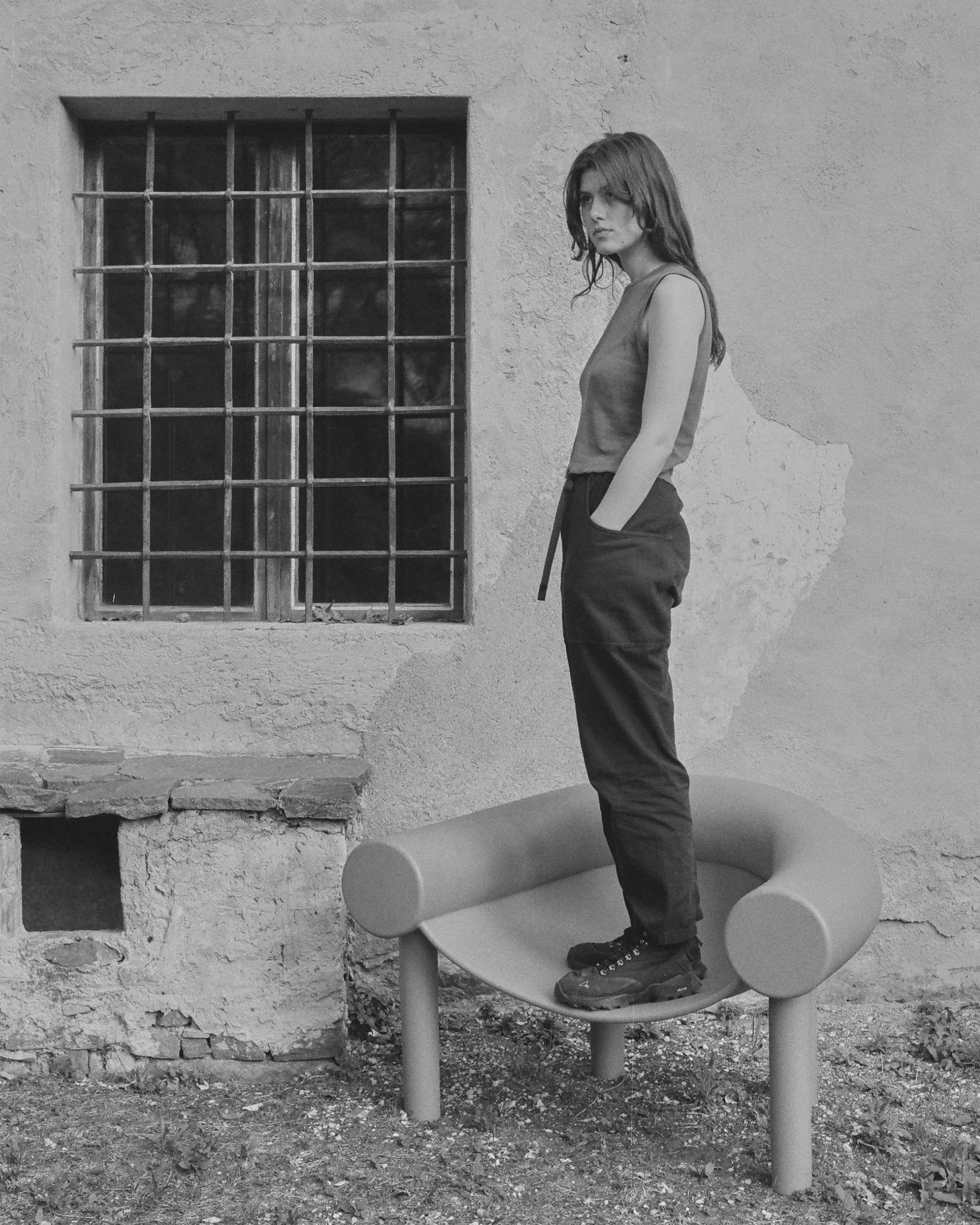 A young woman with long hair standing on a modern, curved bench outdoors, against a textured wall with a barred window, black and white photo.