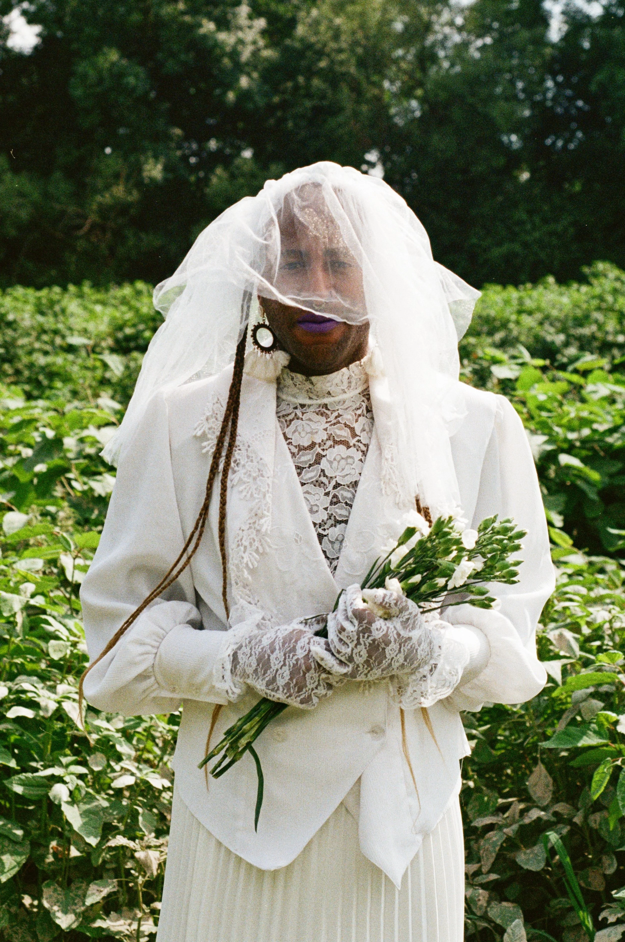 Person dressed in a white lace blazer, gloves, and veil, holding a bouquet of white flowers, standing outdoors in a green field.