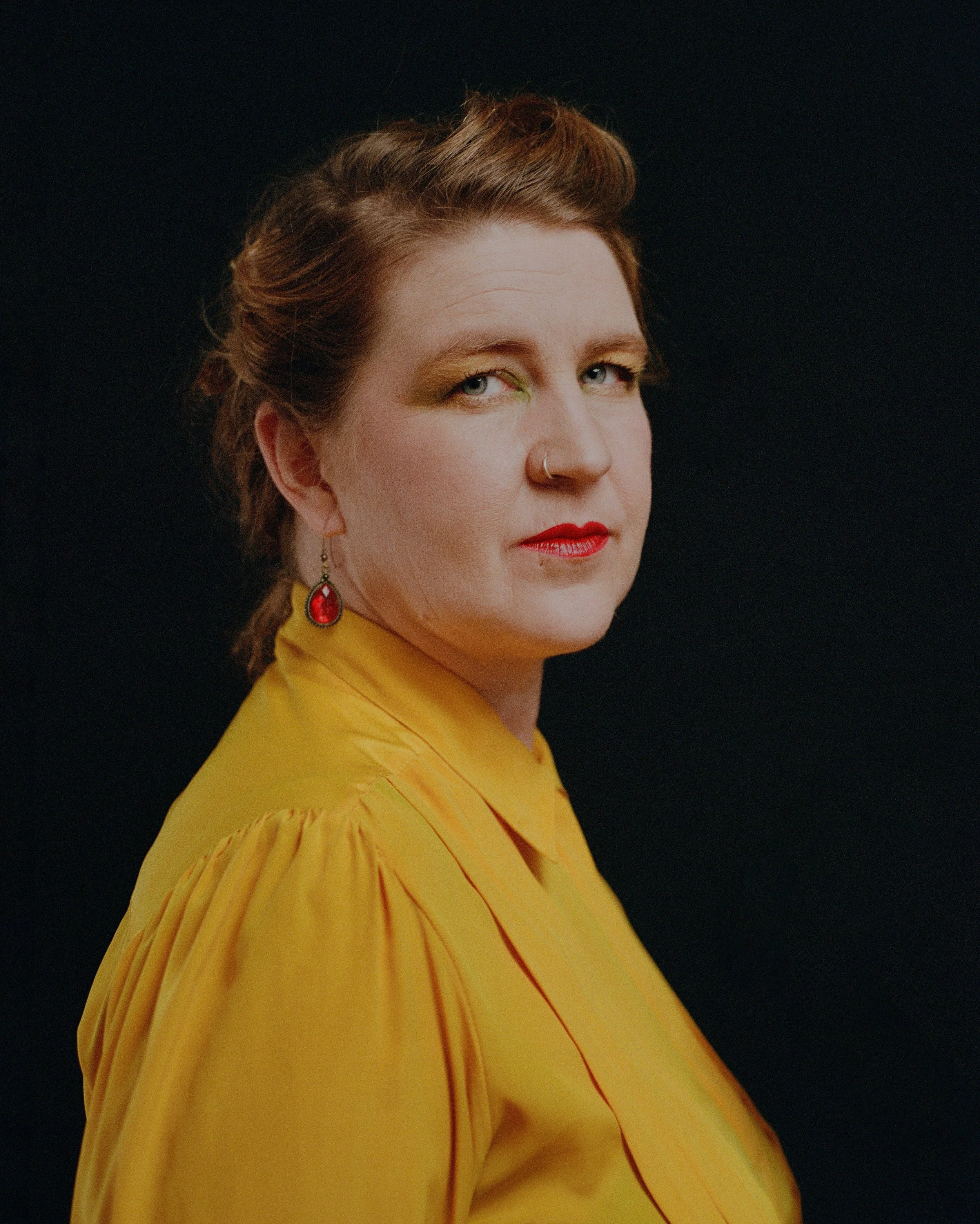 A woman with red hair, light skin, and makeup, wearing a yellow blouse, red earrings, and a nose ring, against a black background.