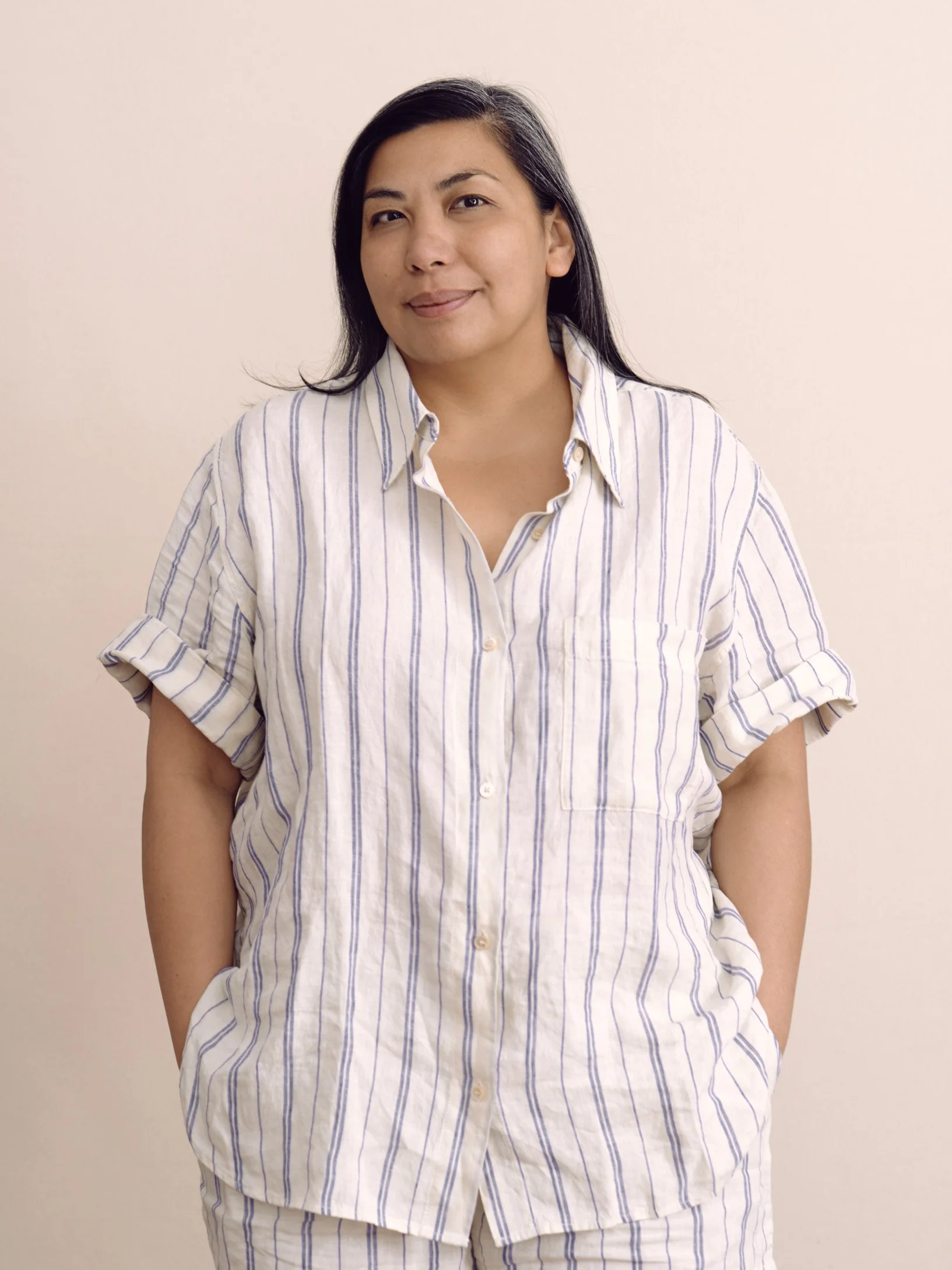 A woman with black hair wearing a cream and blue striped shirt standing against a plain light-colored background.