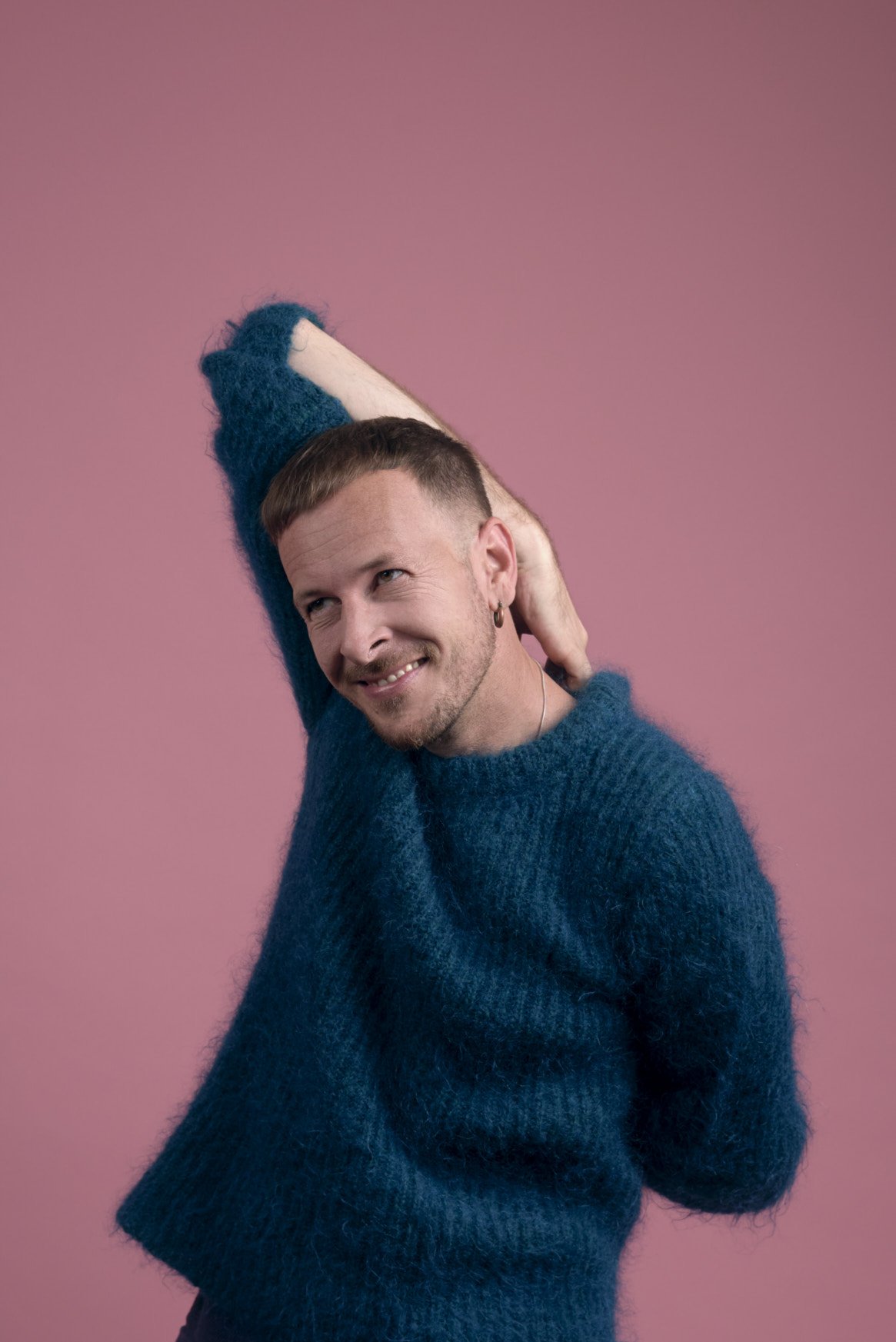 A man with short brown hair and a beard, wearing a blue fuzzy sweater, smiling, with one arm raised behind his head against a pink background.