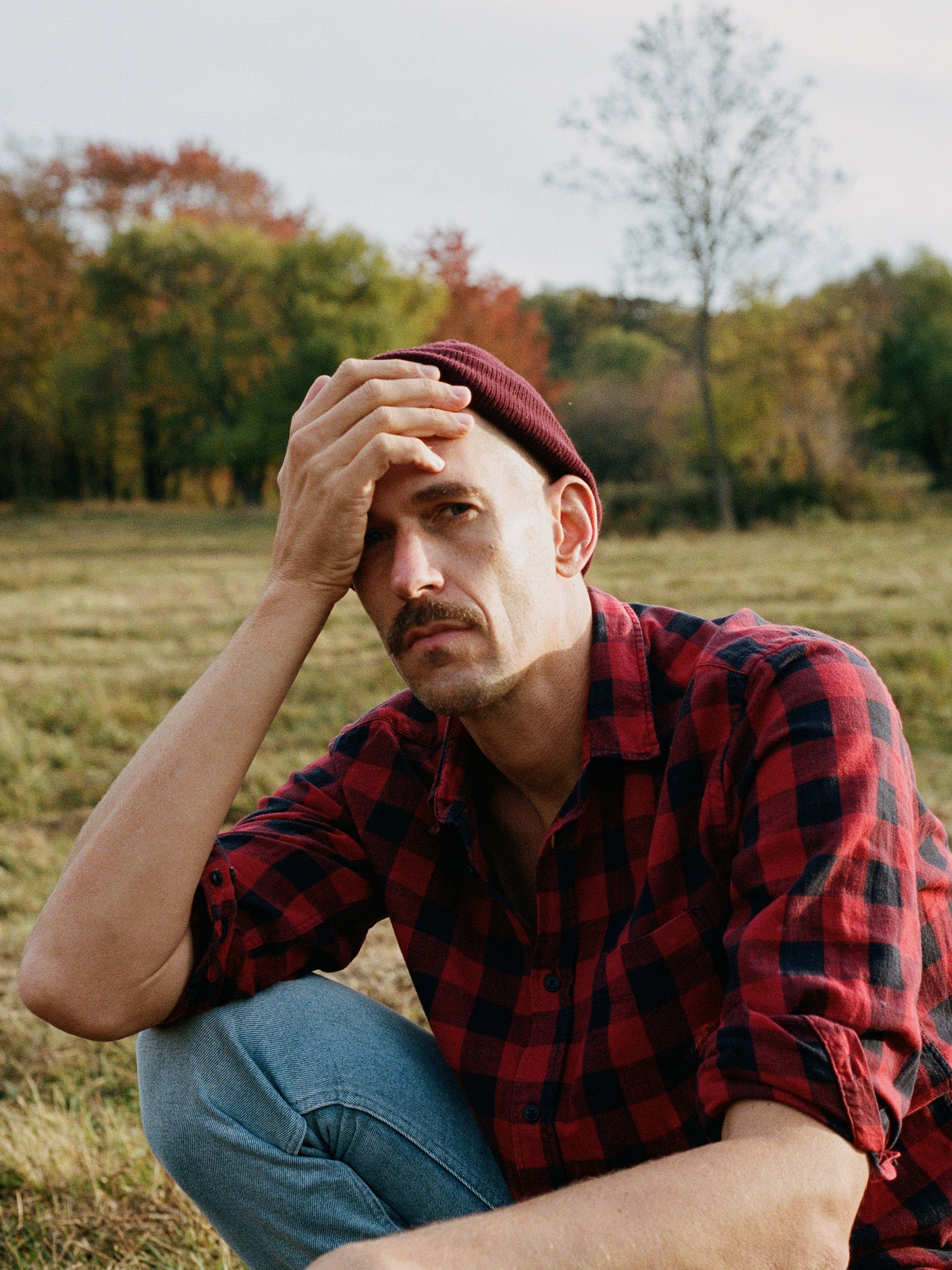 A man with a mustache wearing a maroon beanie and a red and black checkered shirt sitting in a field with autumn trees in the background, holding his forehead with a worried or tired expression.