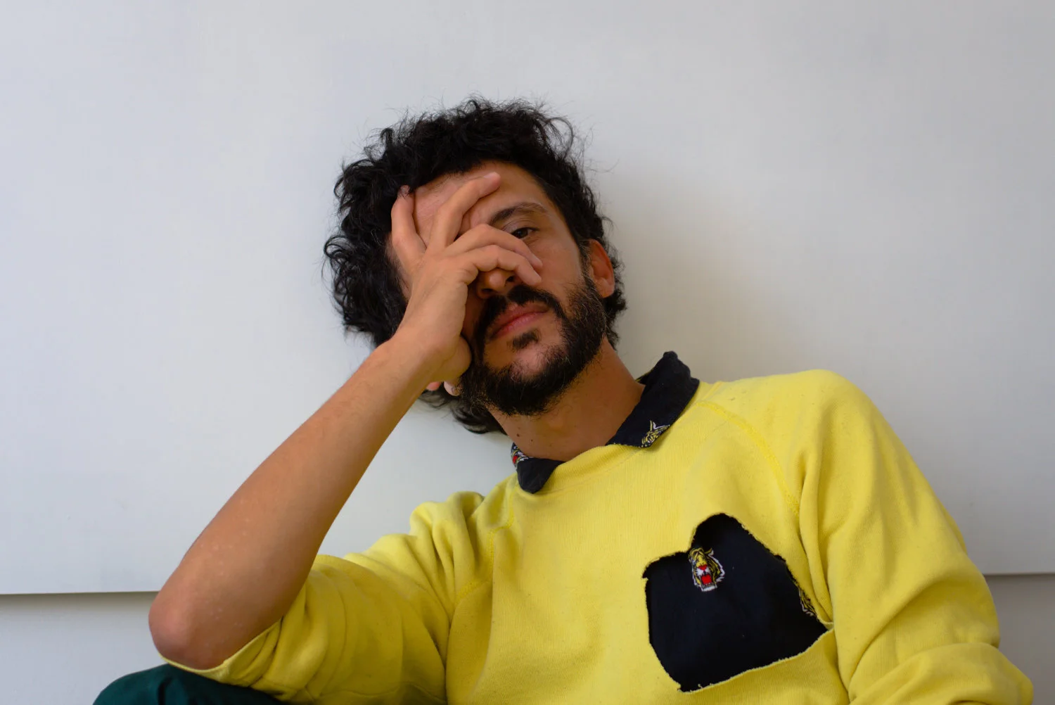 A man with dark curly hair and a beard resting his head on his hand against a plain white wall, wearing a yellow sweatshirt.