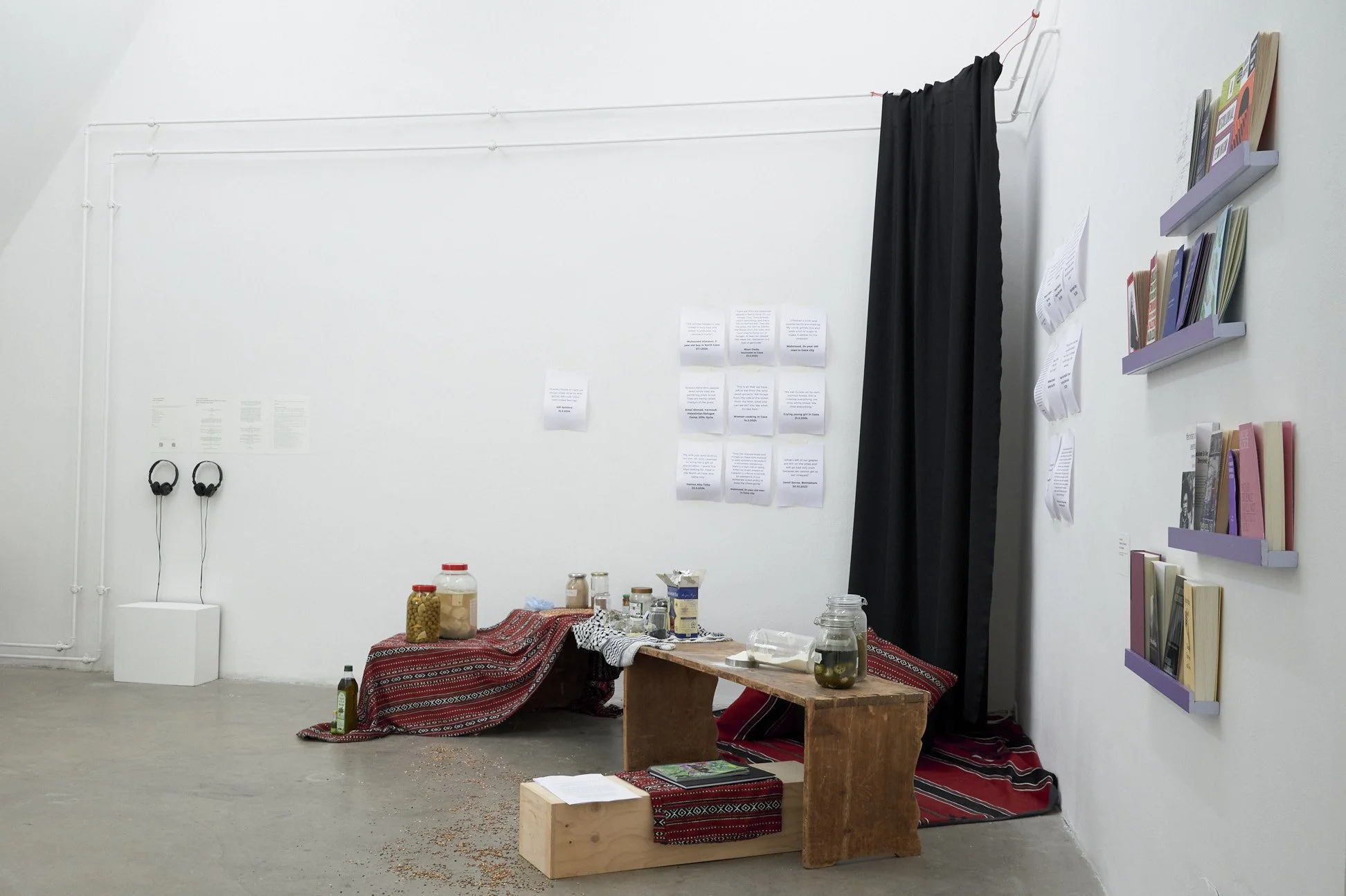 An art installation in a gallery featuring a white wall with hanging black curtain, shelves with books, jars, papers, and a table with textiles and jars, with headphones hanging on the wall.